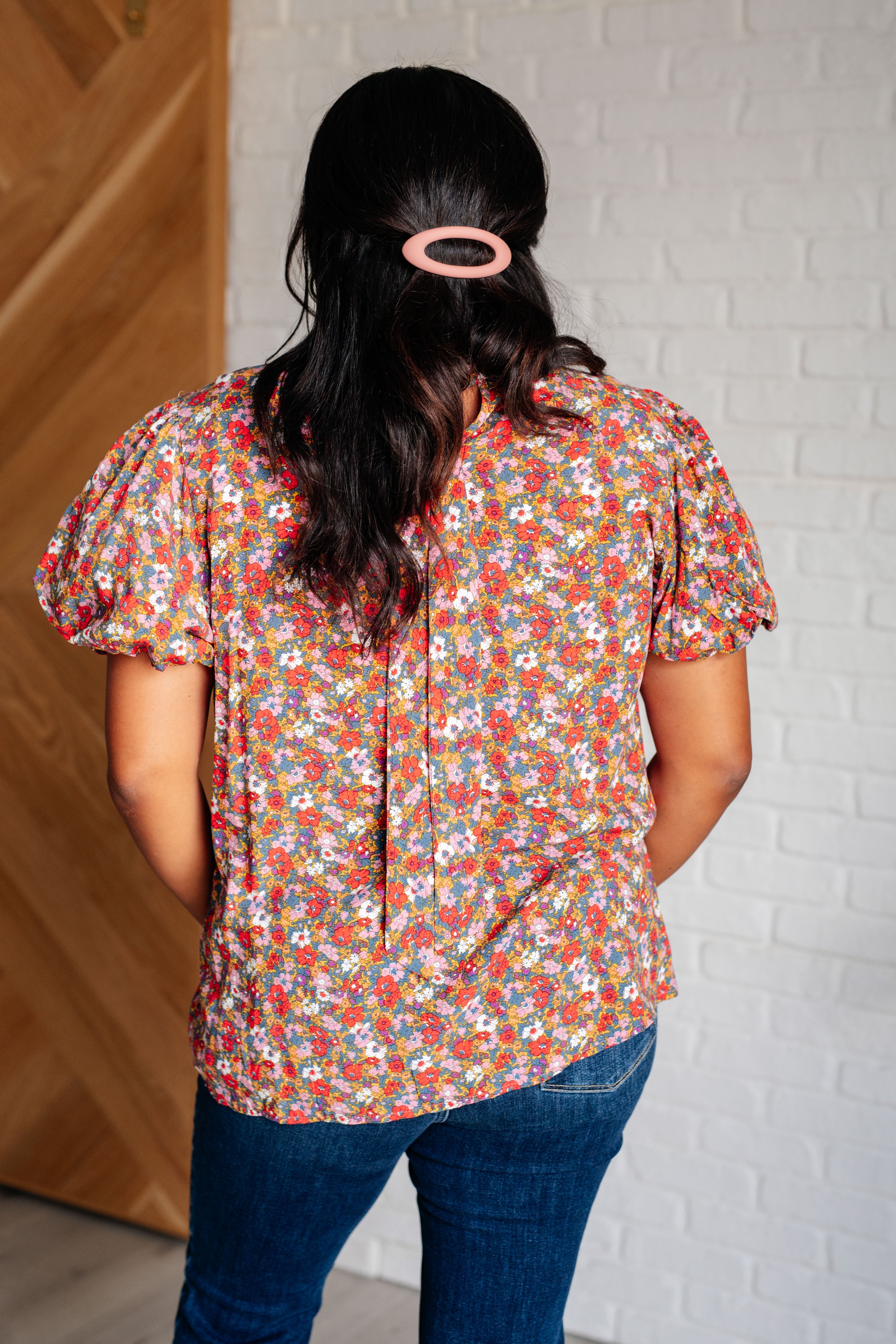 Woman wearing colorful floral puff sleeve blouse with mock neck tie detail and dark jeans standing indoors back view