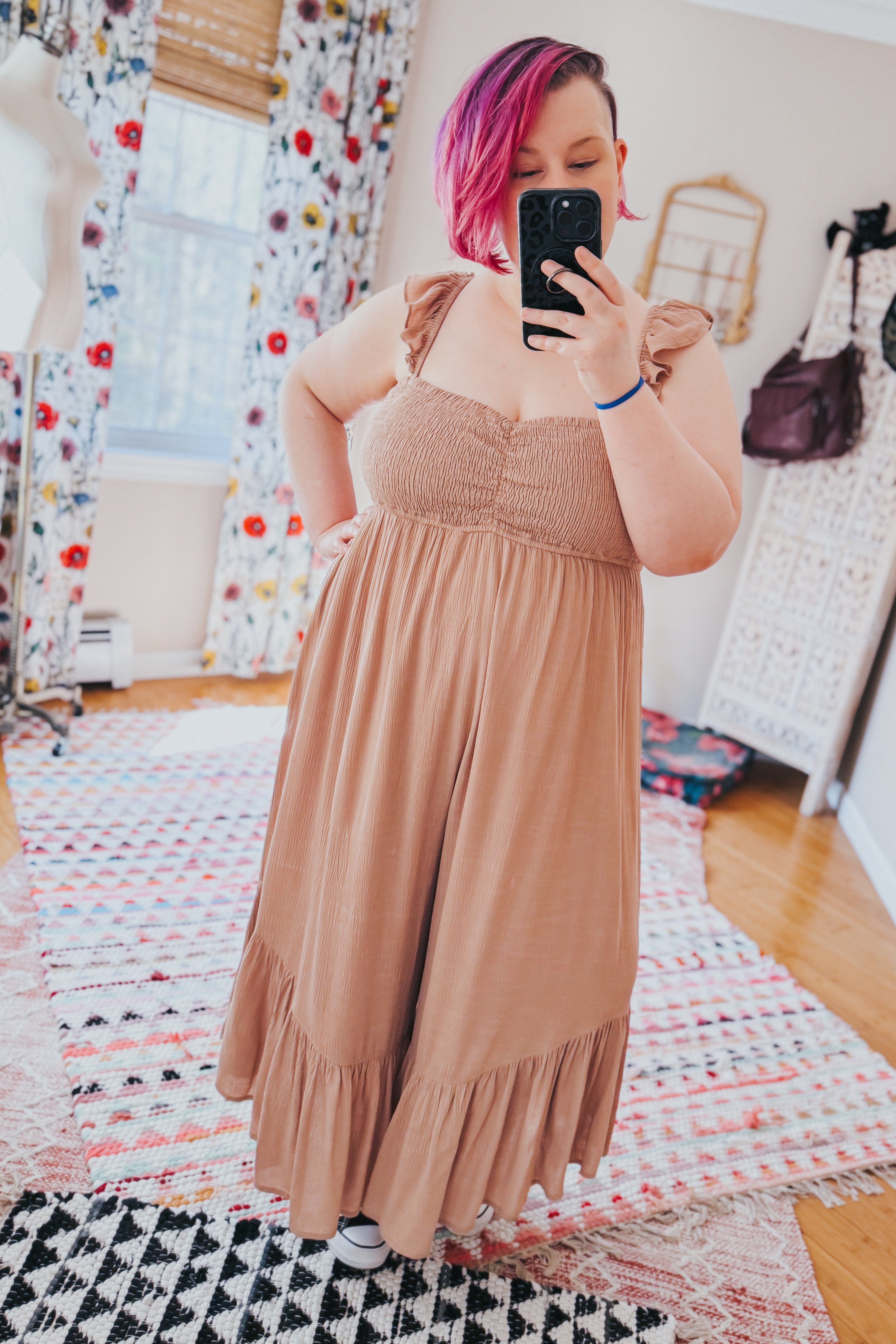 Woman with short pink hair taking selfie wearing light brown wide leg jumpsuit with ruffle straps indoors on patterned rugs