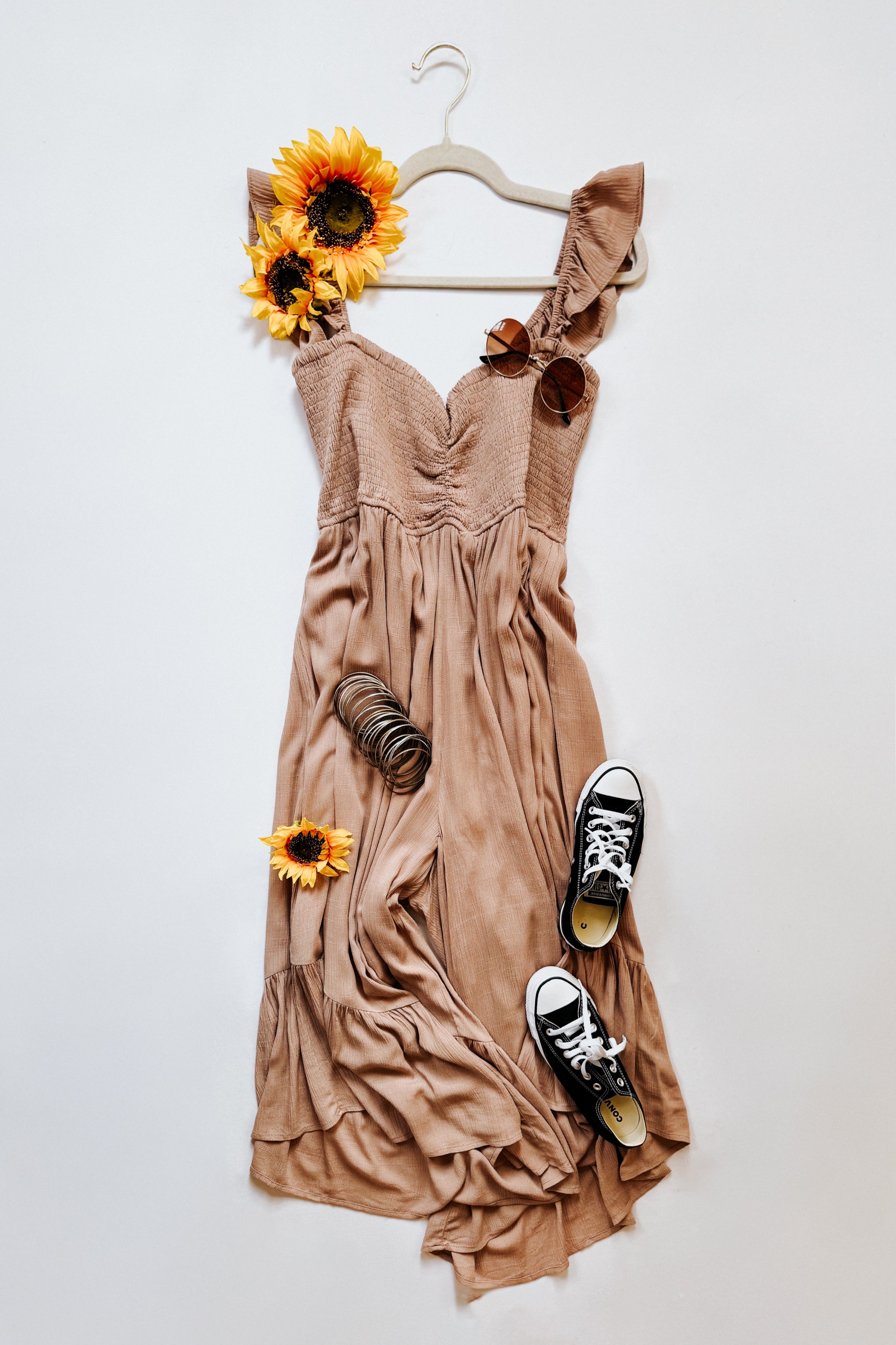 Light brown wide leg jumpsuit with ruched bodice and ruffle shoulder strap styled with sunglasses and sneakers on white background