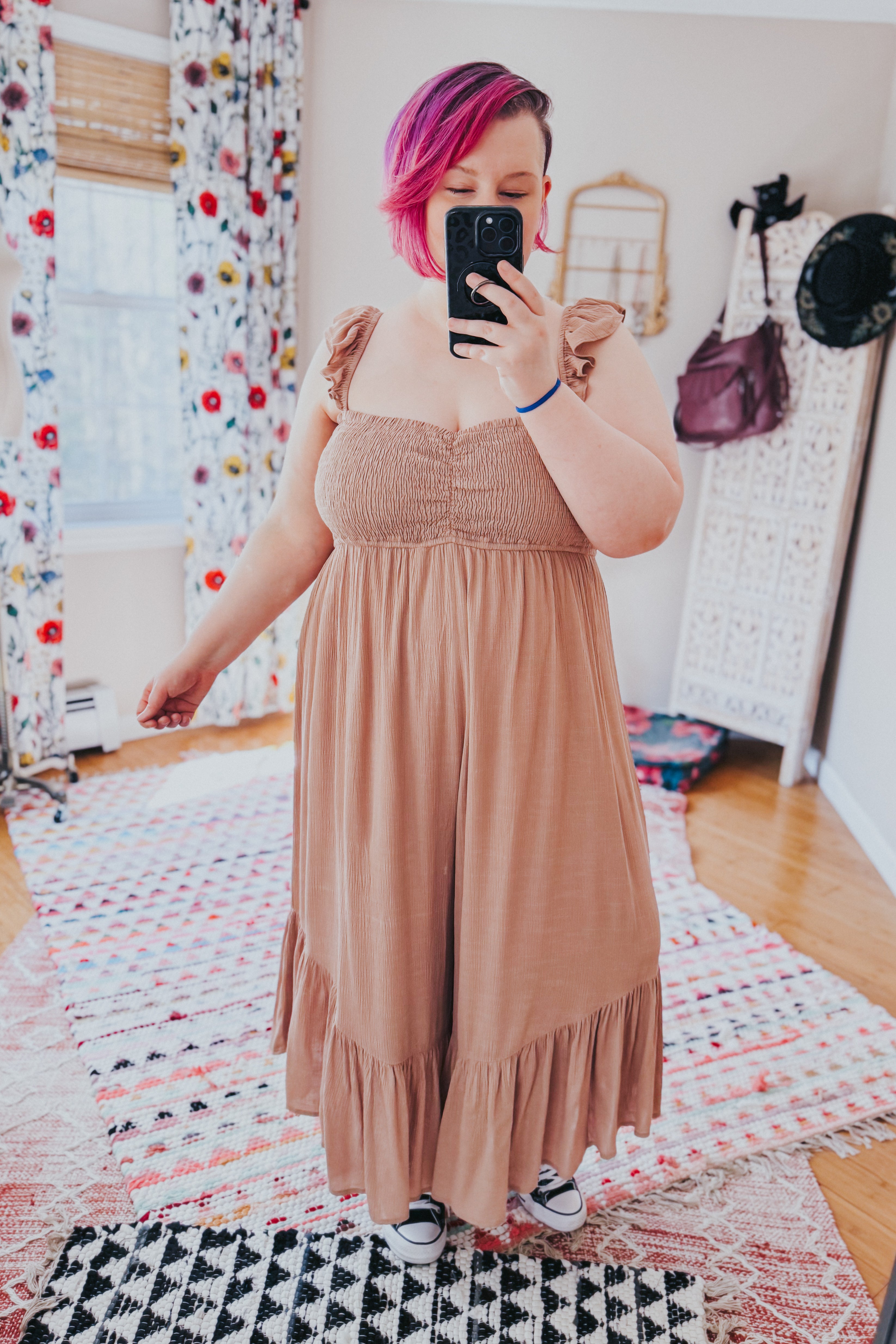 Woman with pink hair taking mirror selfie wearing light brown smocked bodice wide leg jumpsuit with ruffle shoulder straps indoors