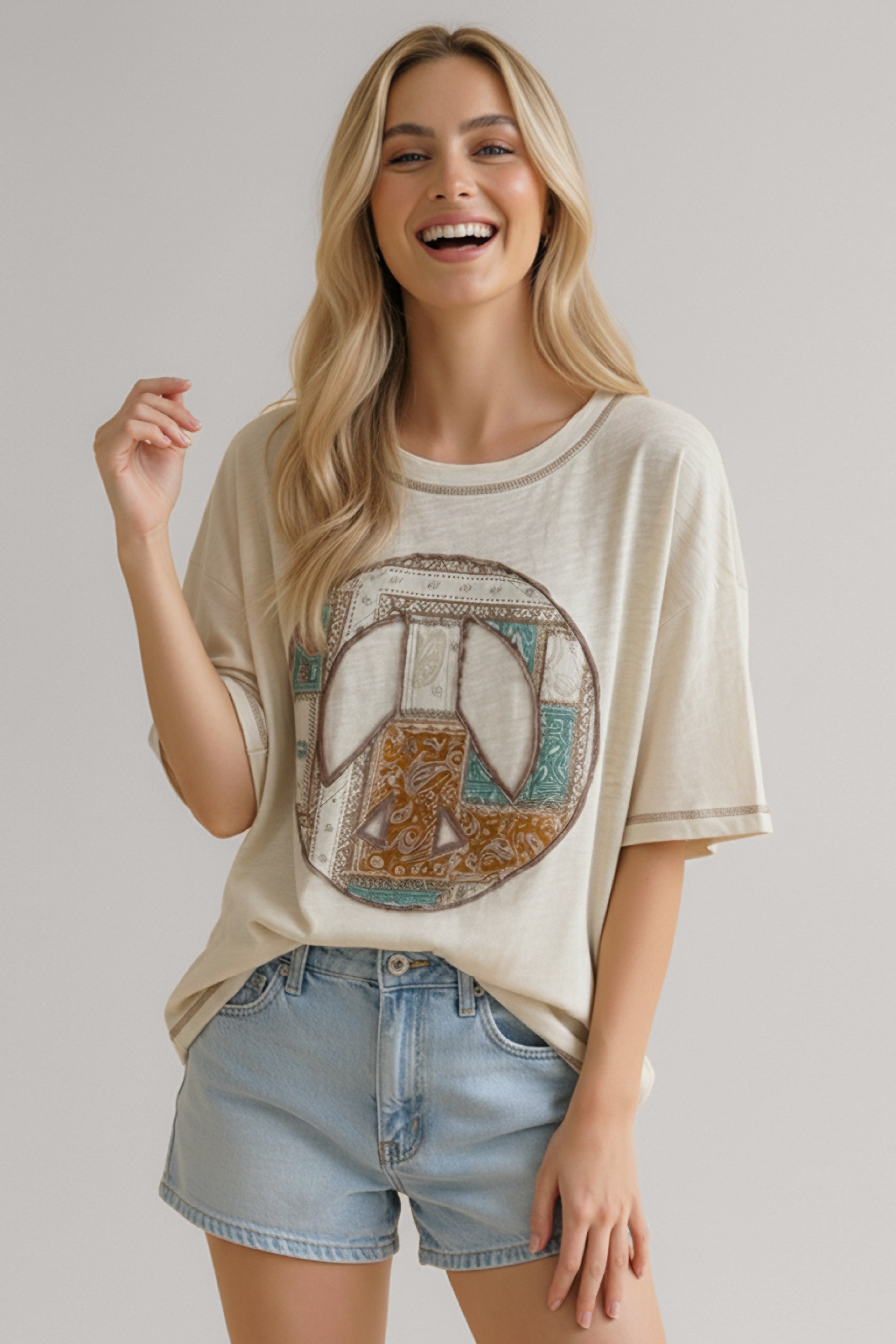 Smiling woman wearing a cream loose-fit tee with a multicolor patchwork peace sign graphic and light denim shorts