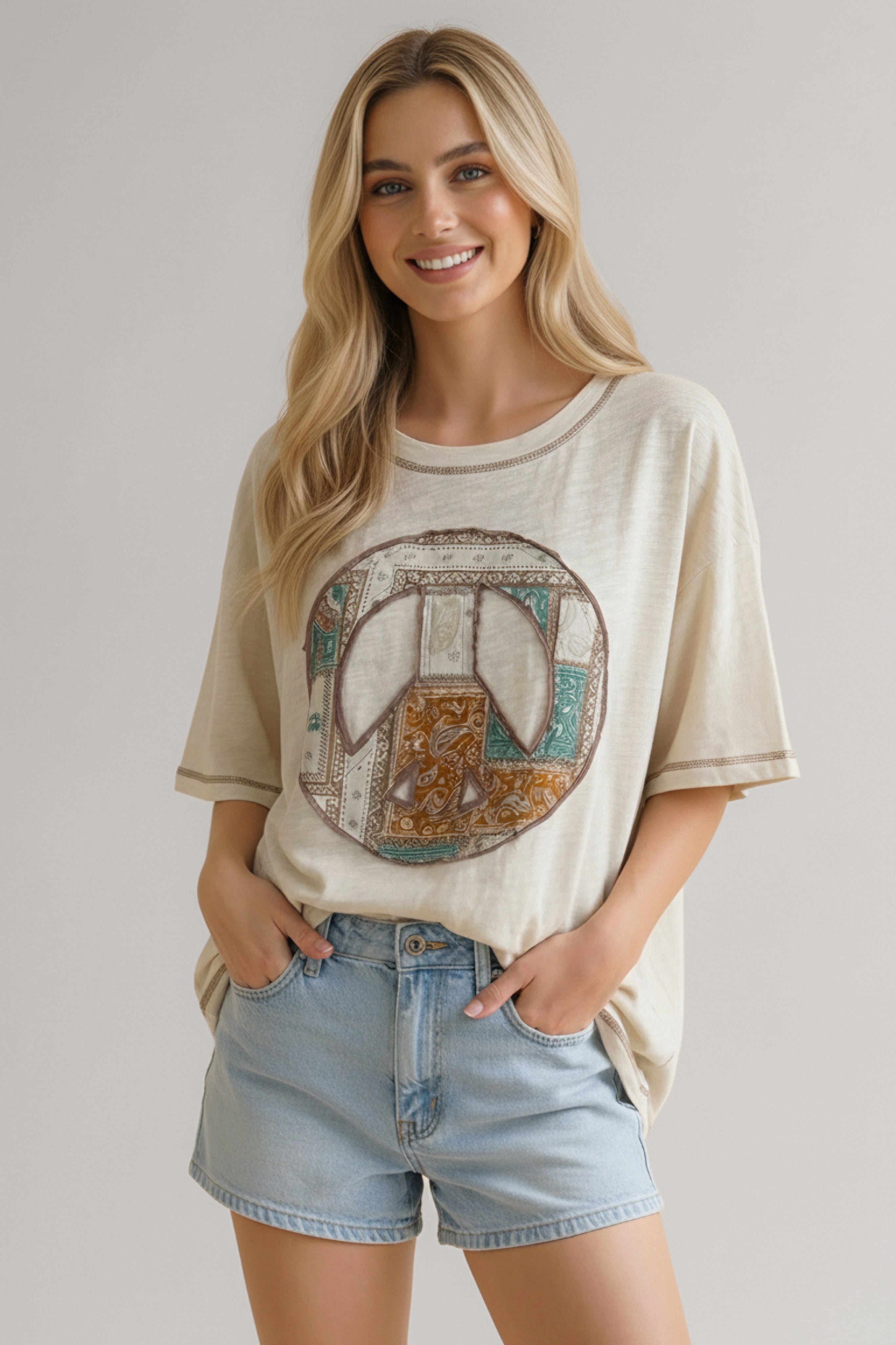 Smiling woman wearing a cream oversized tee with a multicolor patterned peace sign graphic and light denim shorts