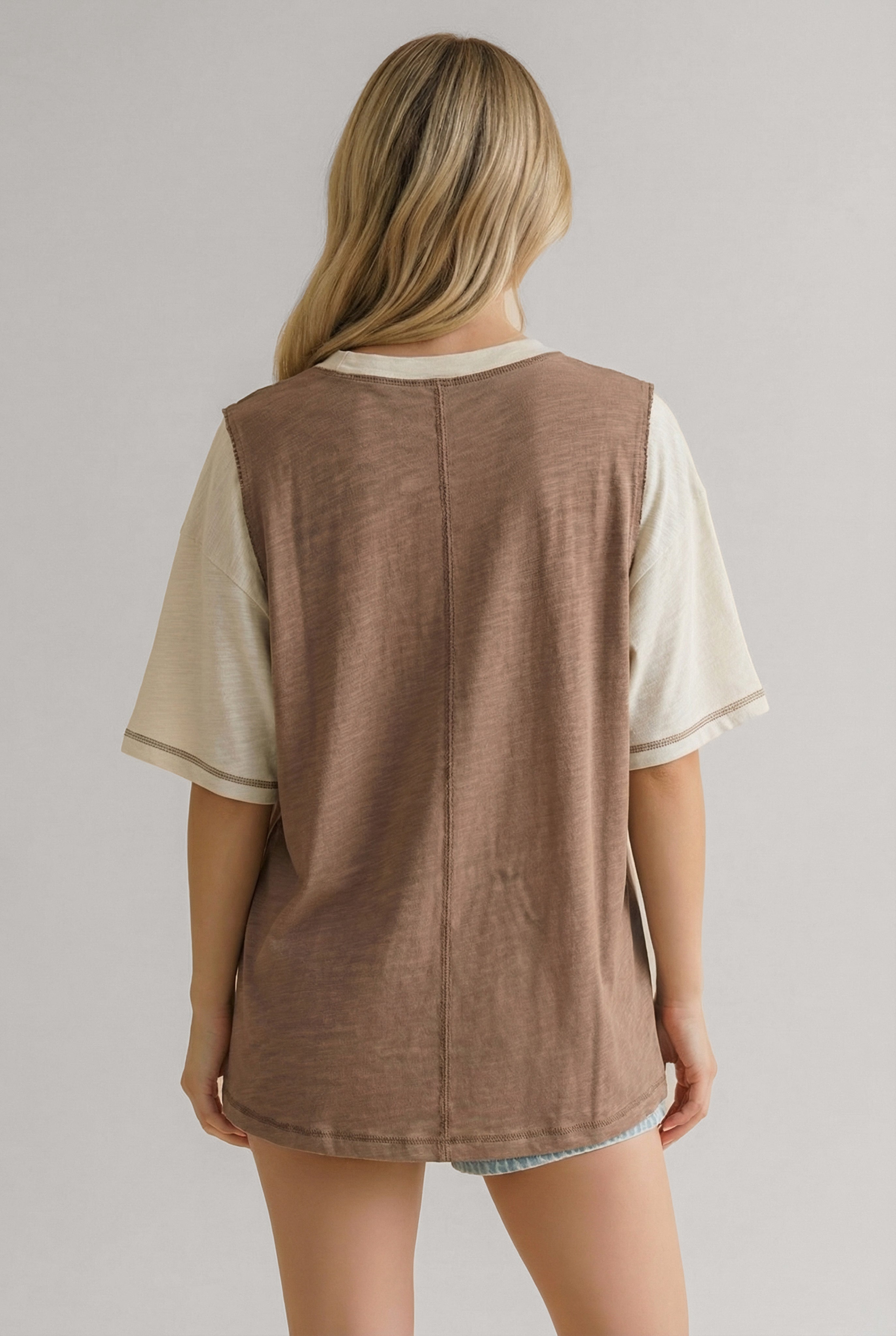 Woman with long blonde hair wearing a cream short-sleeve tee layered under a sleeveless brown top viewed from the back