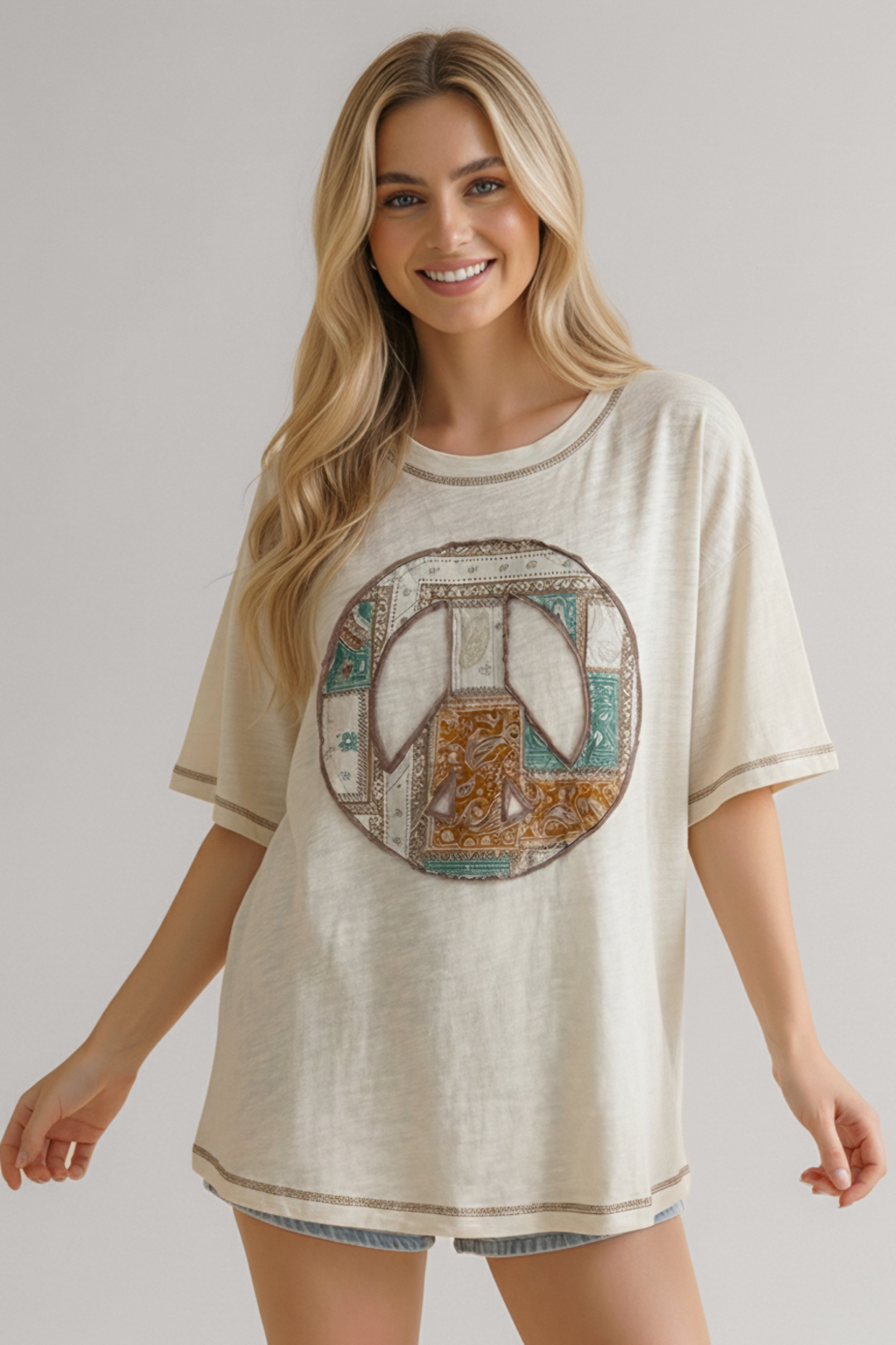 Smiling woman wearing a cream oversized tee with a multicolor patchwork peace sign graphic and light blue denim shorts