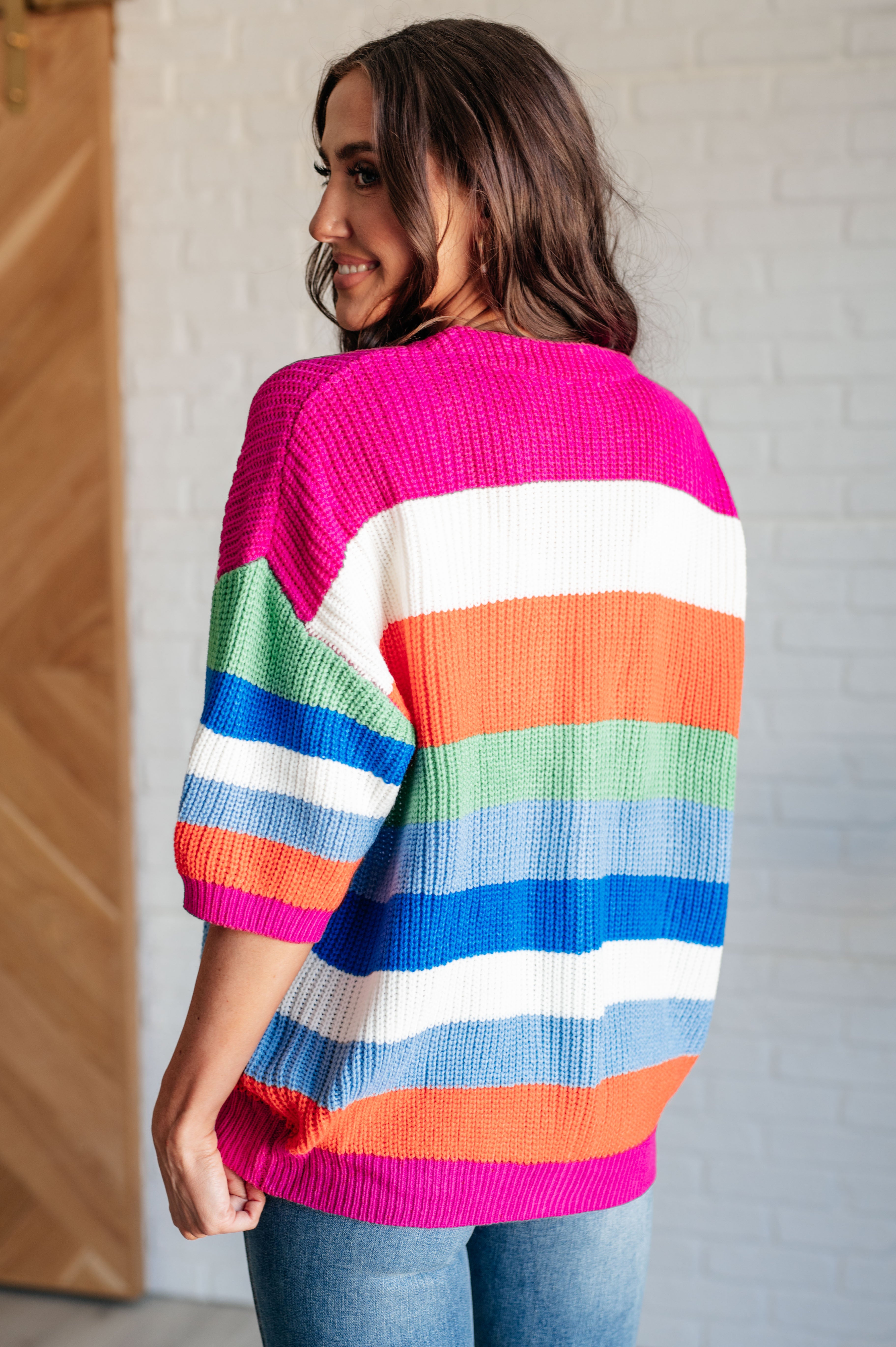 Woman wearing vibrant striped knit cardigan in pink, orange, green, blue, and white, shown from the back with jeans