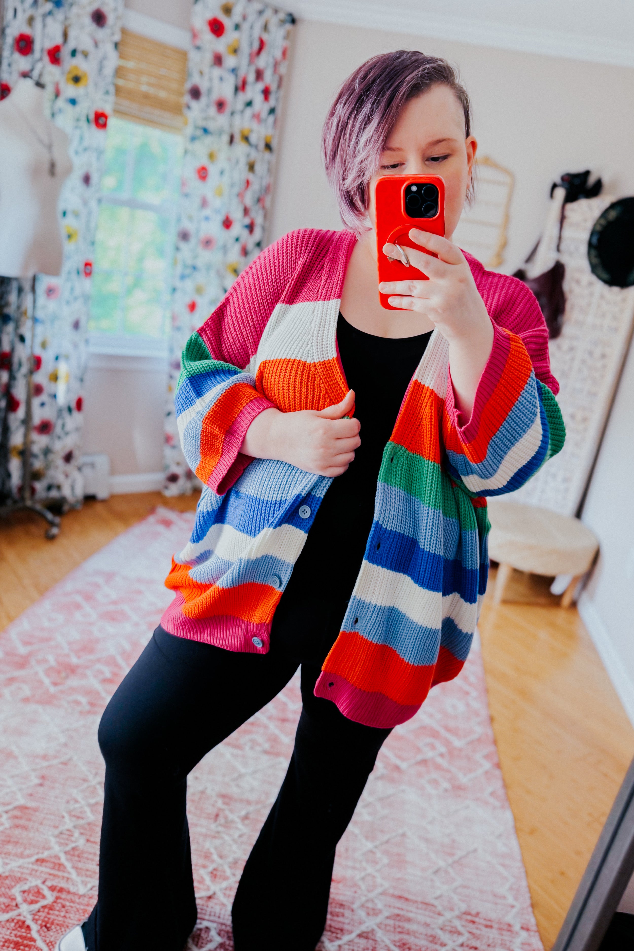 Woman taking mirror selfie wearing colorful striped button down cardigan with black outfit indoors