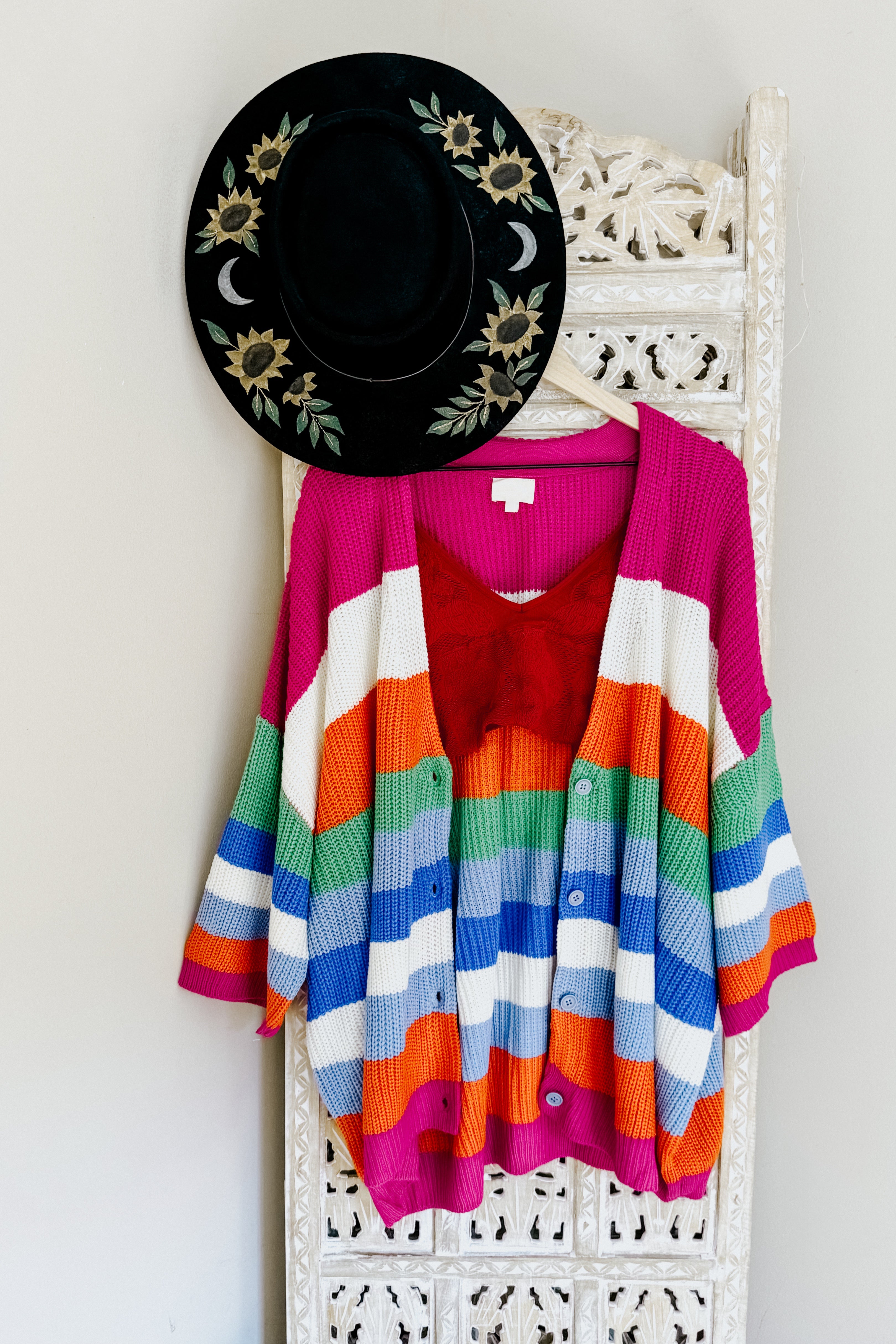Bright multicolored striped knit cardigan paired with a red top hanging on white carved wooden panel with black floral hat