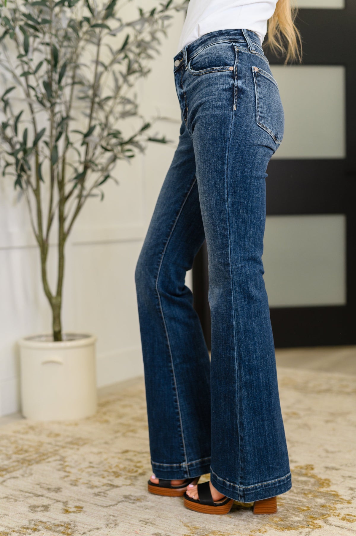 Side view of a woman wearing blue high waist flare jeans with trouser styling and heeled shoes in a bright indoor setting