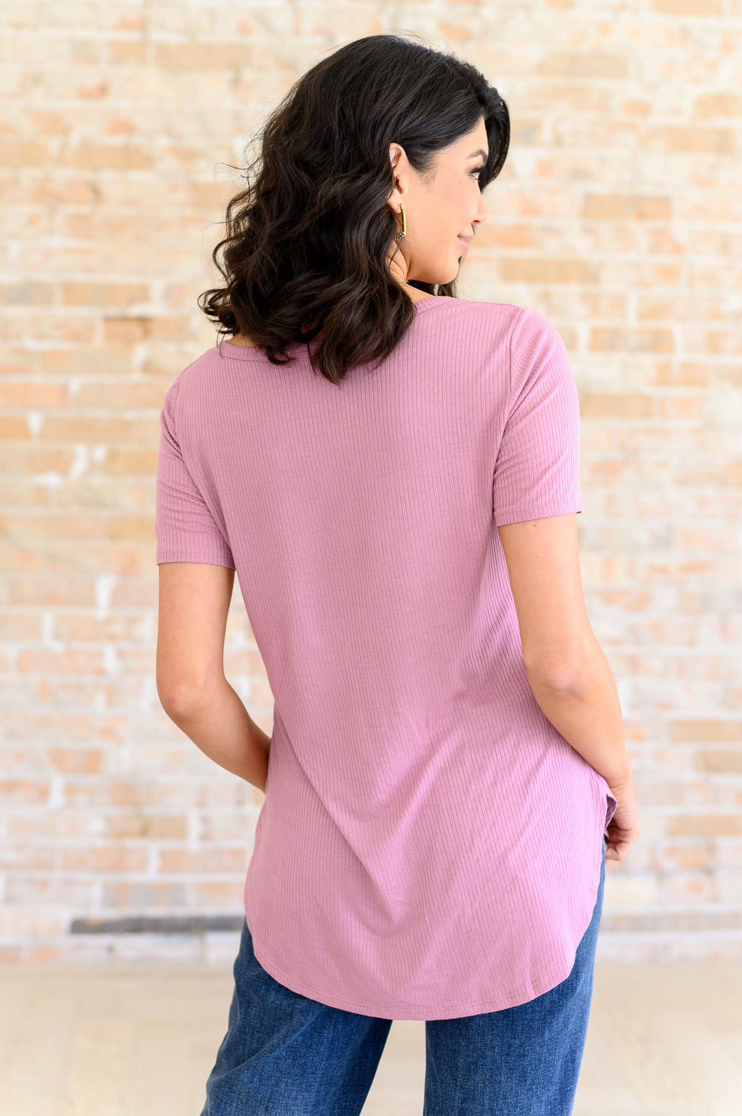 Woman wearing pink ribbed v-neck top with short sleeves and high-low hem standing against brick wall