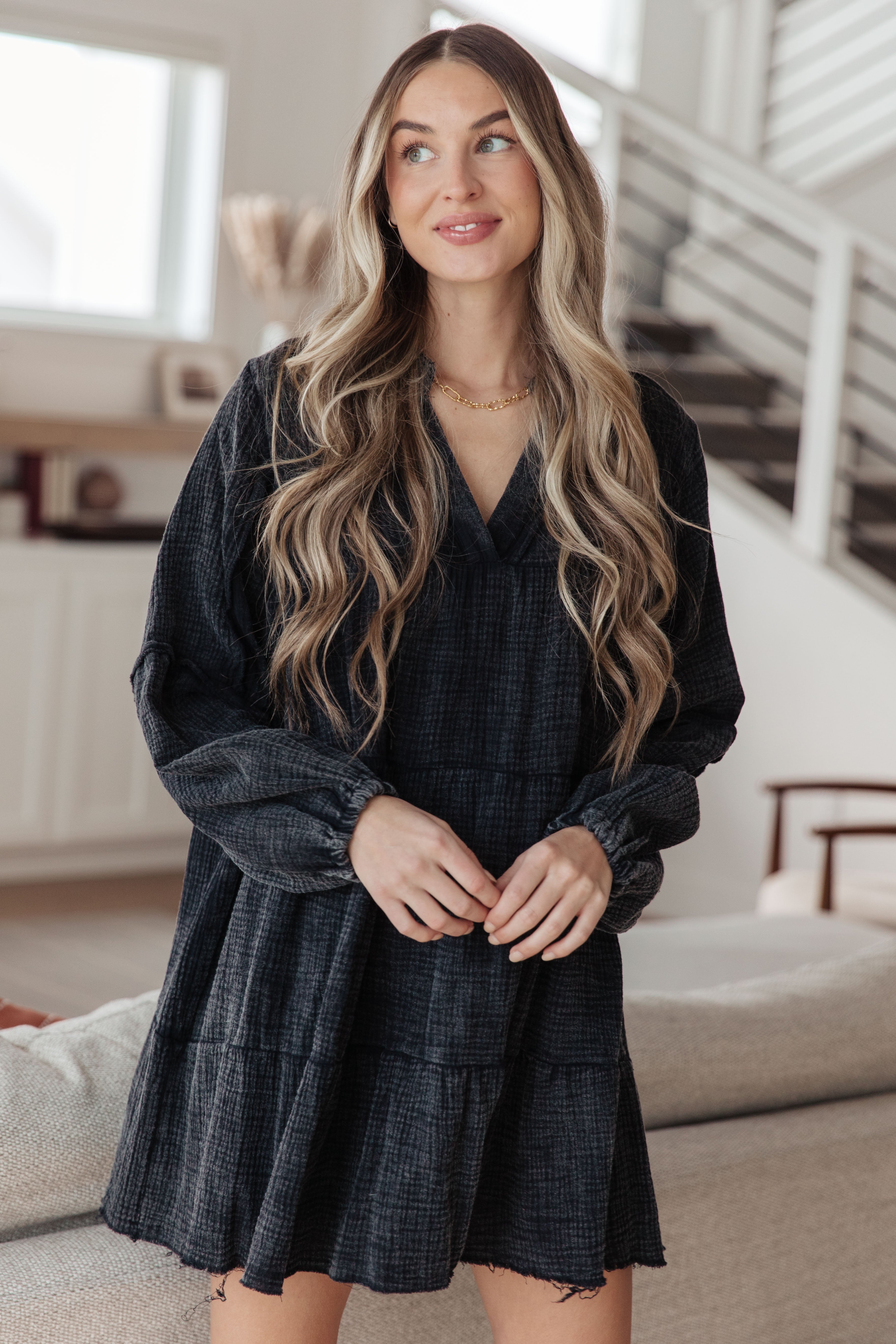 Woman with long wavy blonde hair wearing loose textured black mineral wash V-neck dress indoors