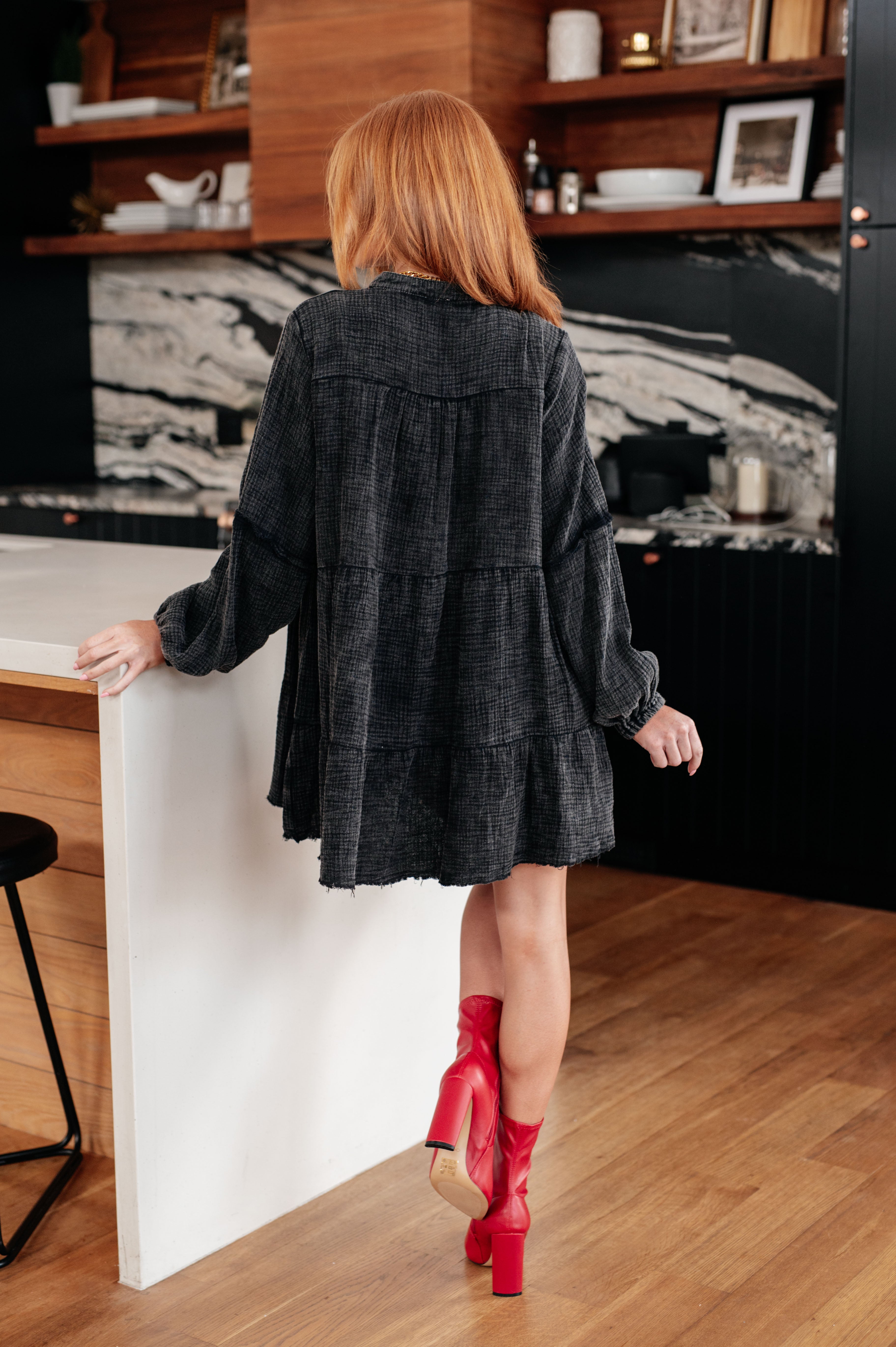 Woman with red hair wearing loose dark mineral wash tiered dress and red heeled ankle boots indoors