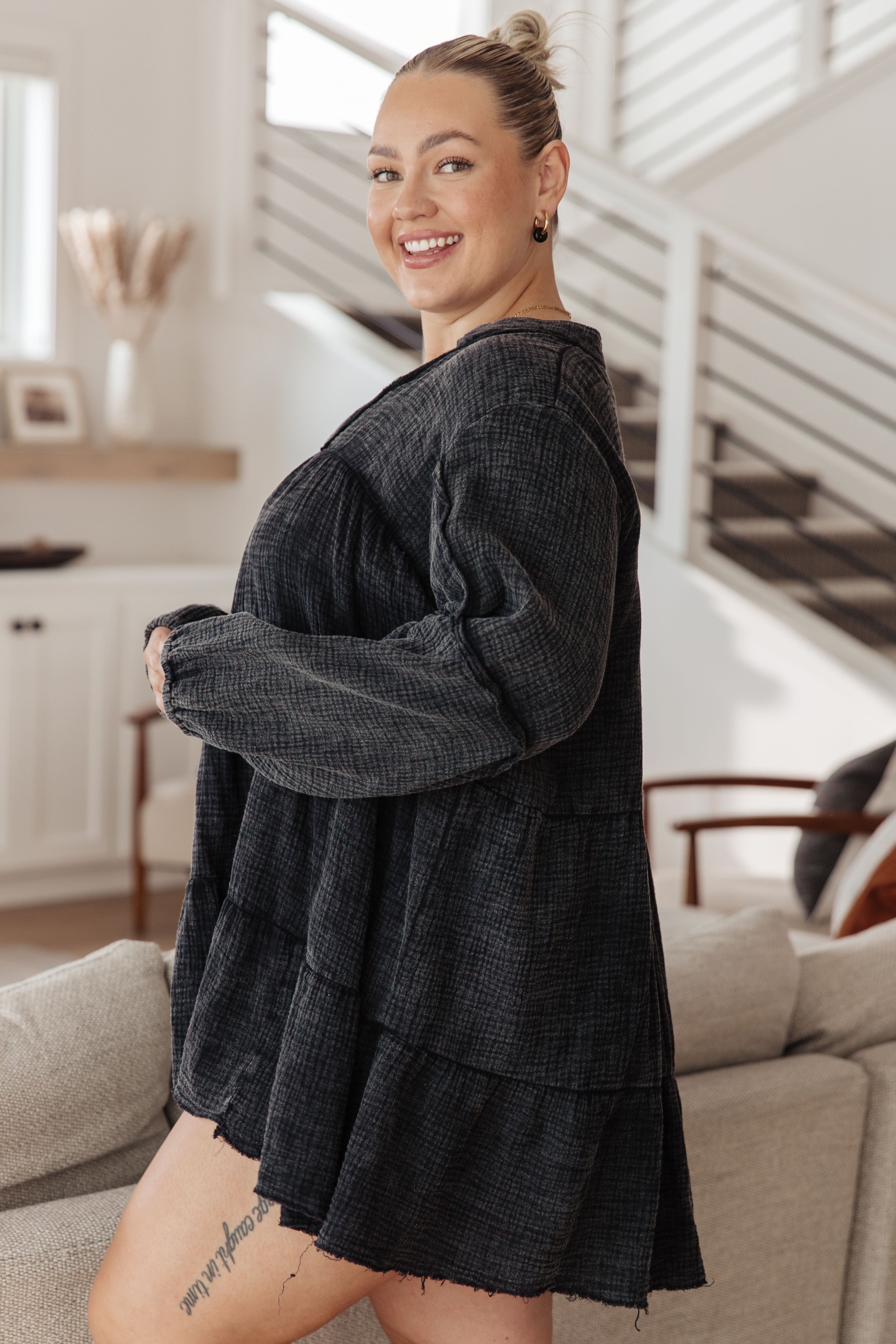 Smiling woman wearing loose dark mineral wash V-neck dress with balloon sleeves standing indoors by sofa