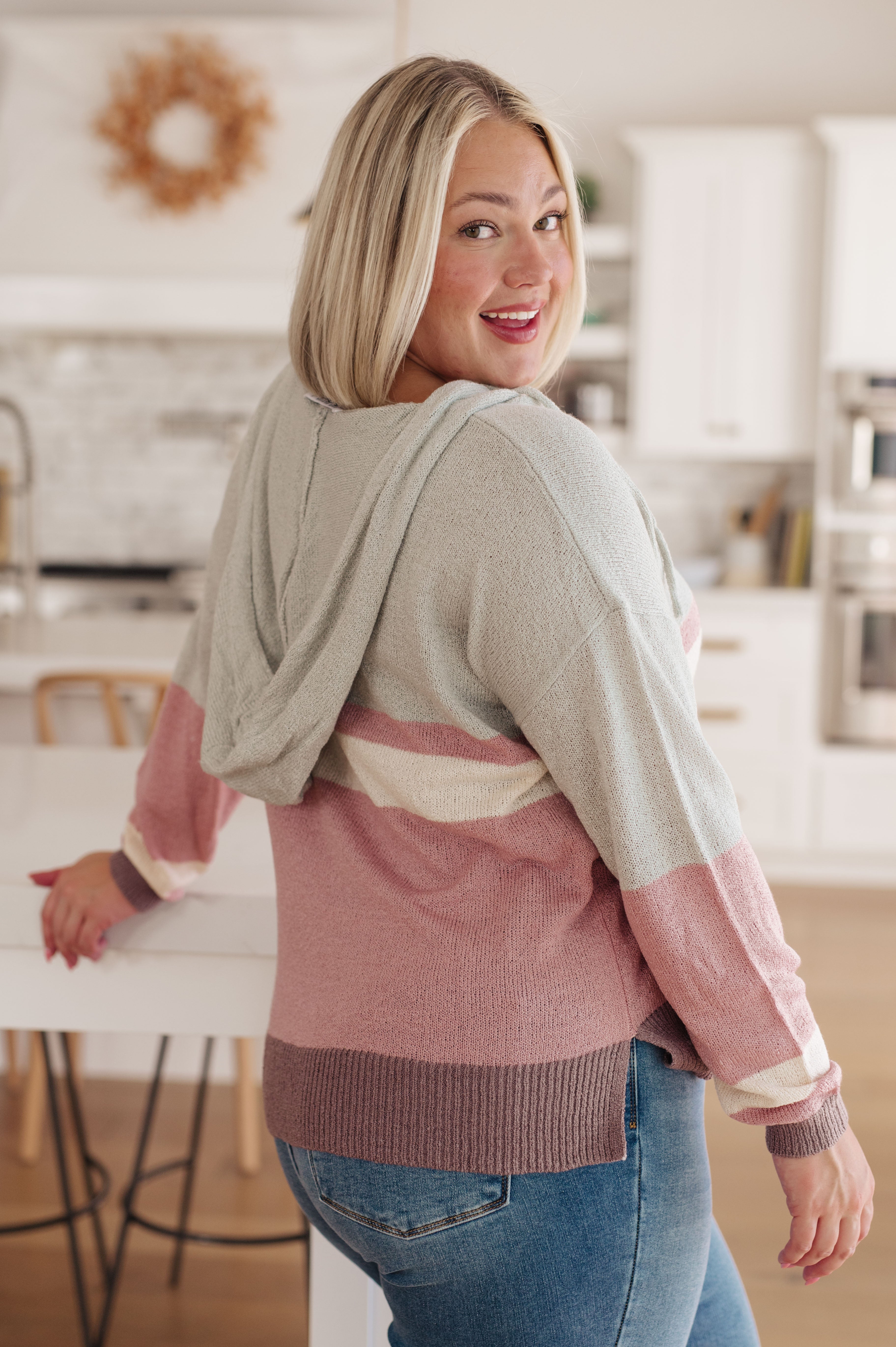 Blonde woman smiling over shoulder wearing pastel color-blocked Boy Problem Spring Hoodie in bright kitchen