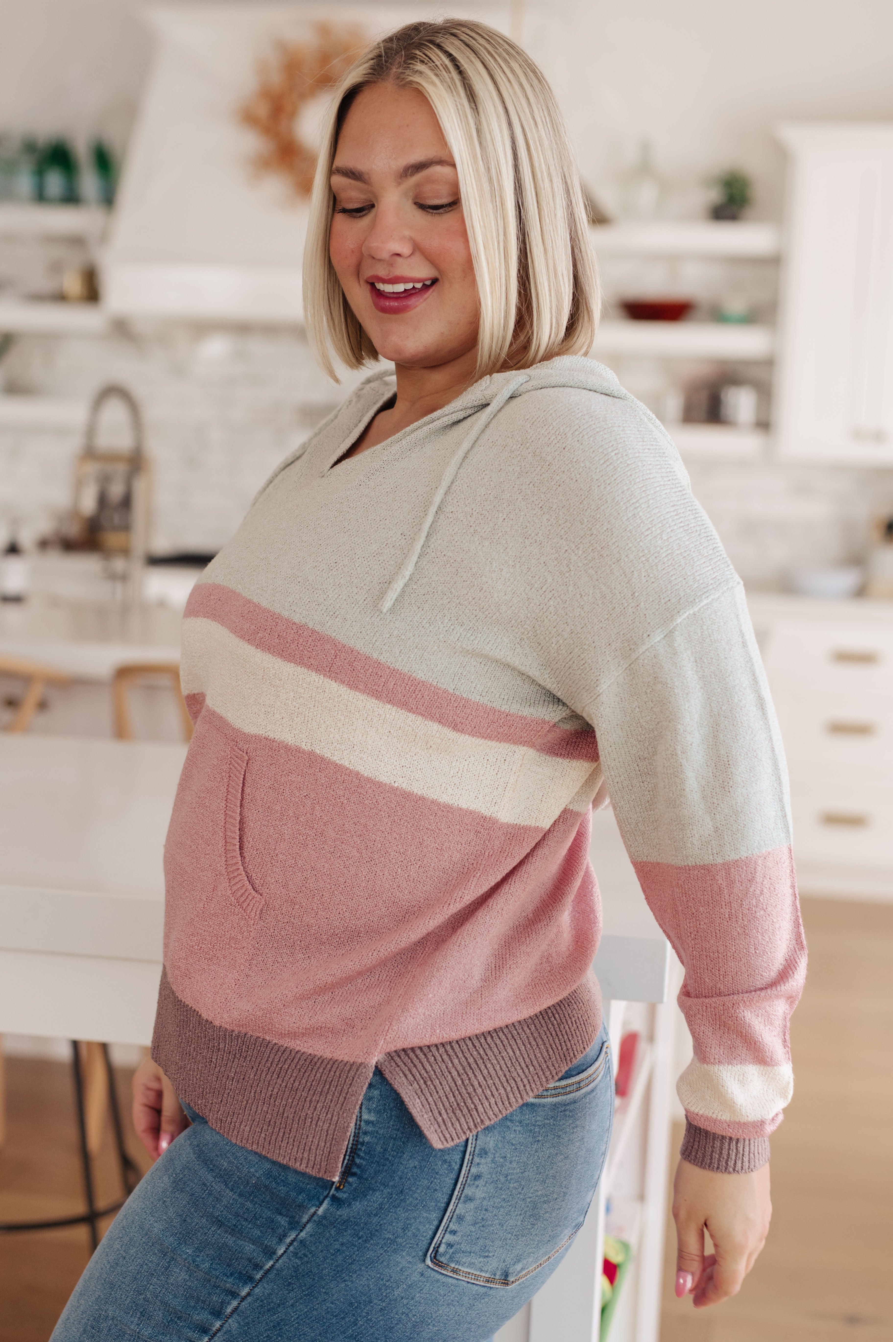 Smiling woman wearing color-blocked knit hoodie with pink, cream, and mauve stripes standing in bright kitchen