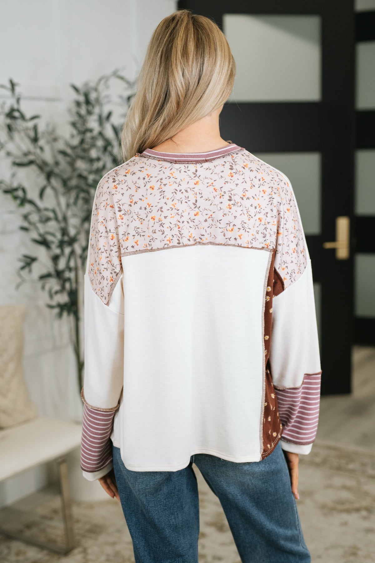 Back view of a woman wearing a cream and rust color block top with a floral yoke and striped cuffs styled with blue jeans indoors