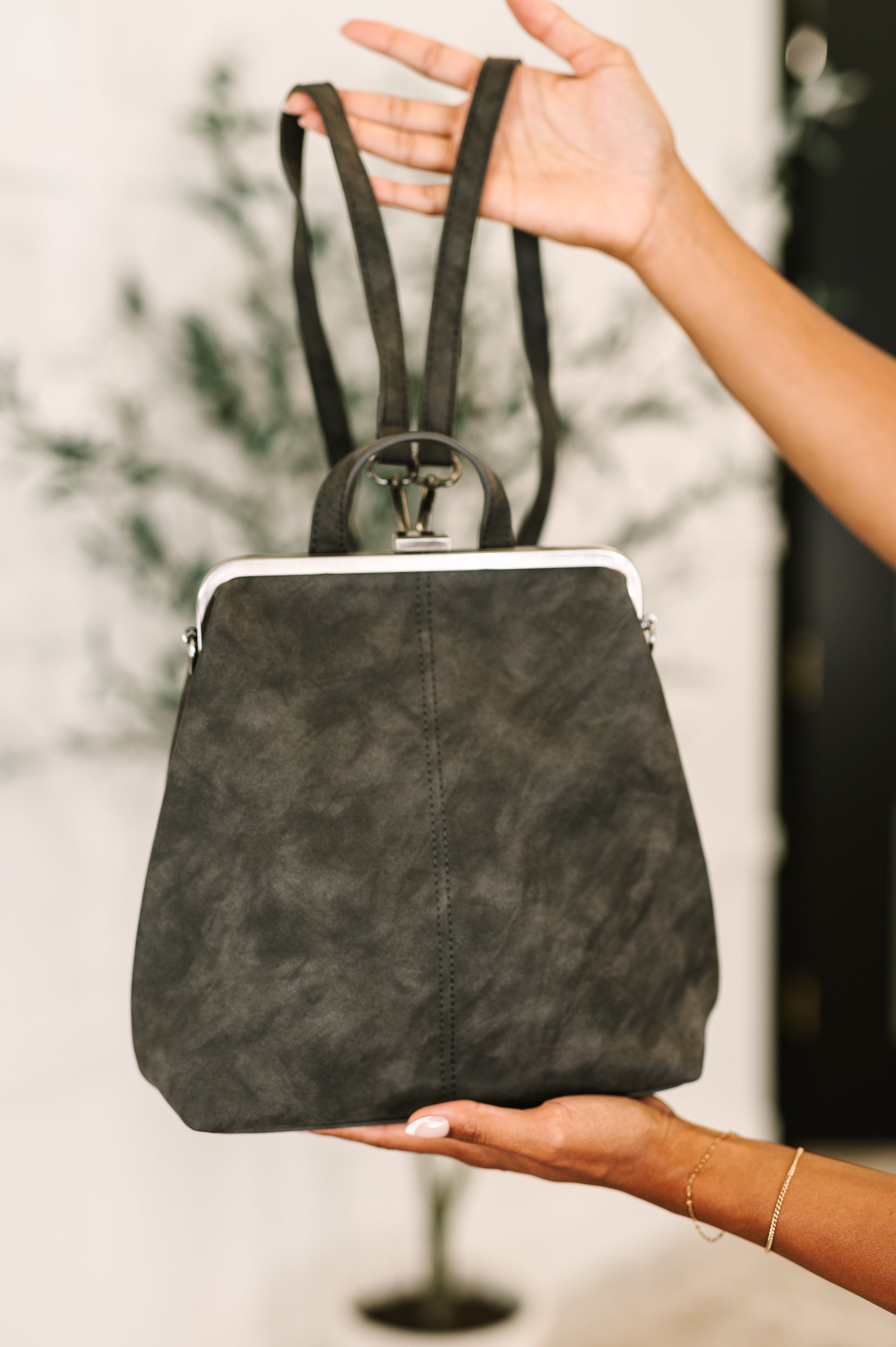 Hands holding a charcoal small backpack with silver frame and soft texture against blurred indoor background