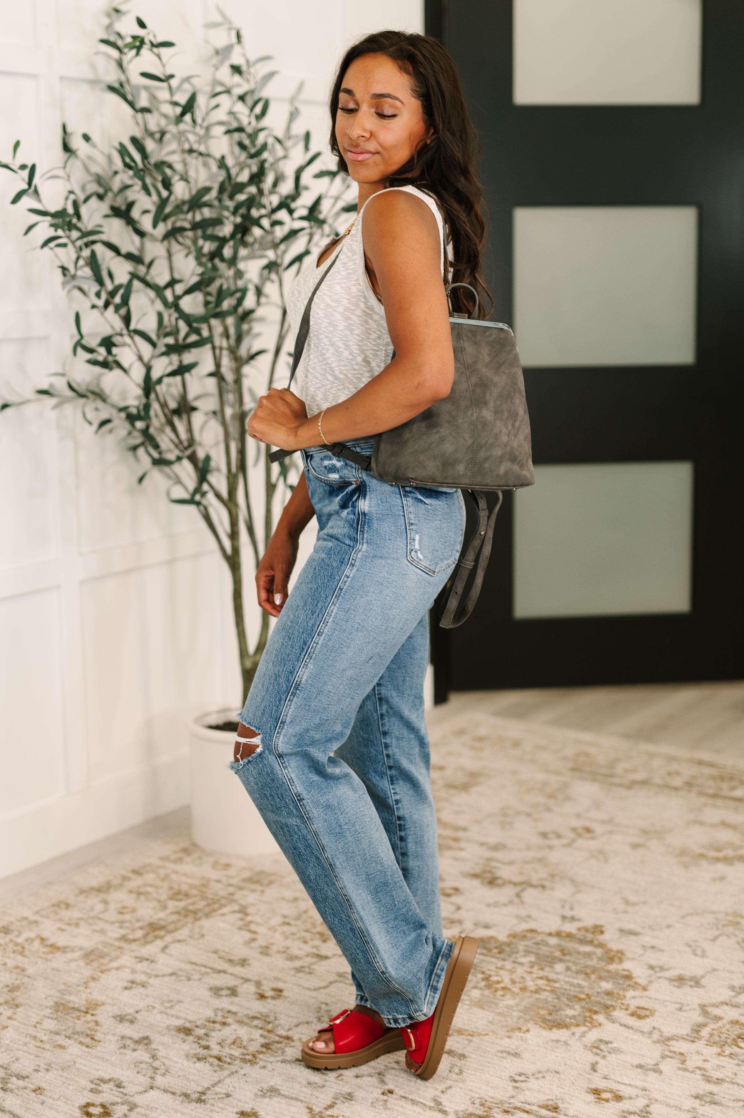 Woman wearing light wash distressed jeans and sleeveless top carrying a charcoal small backpack indoors