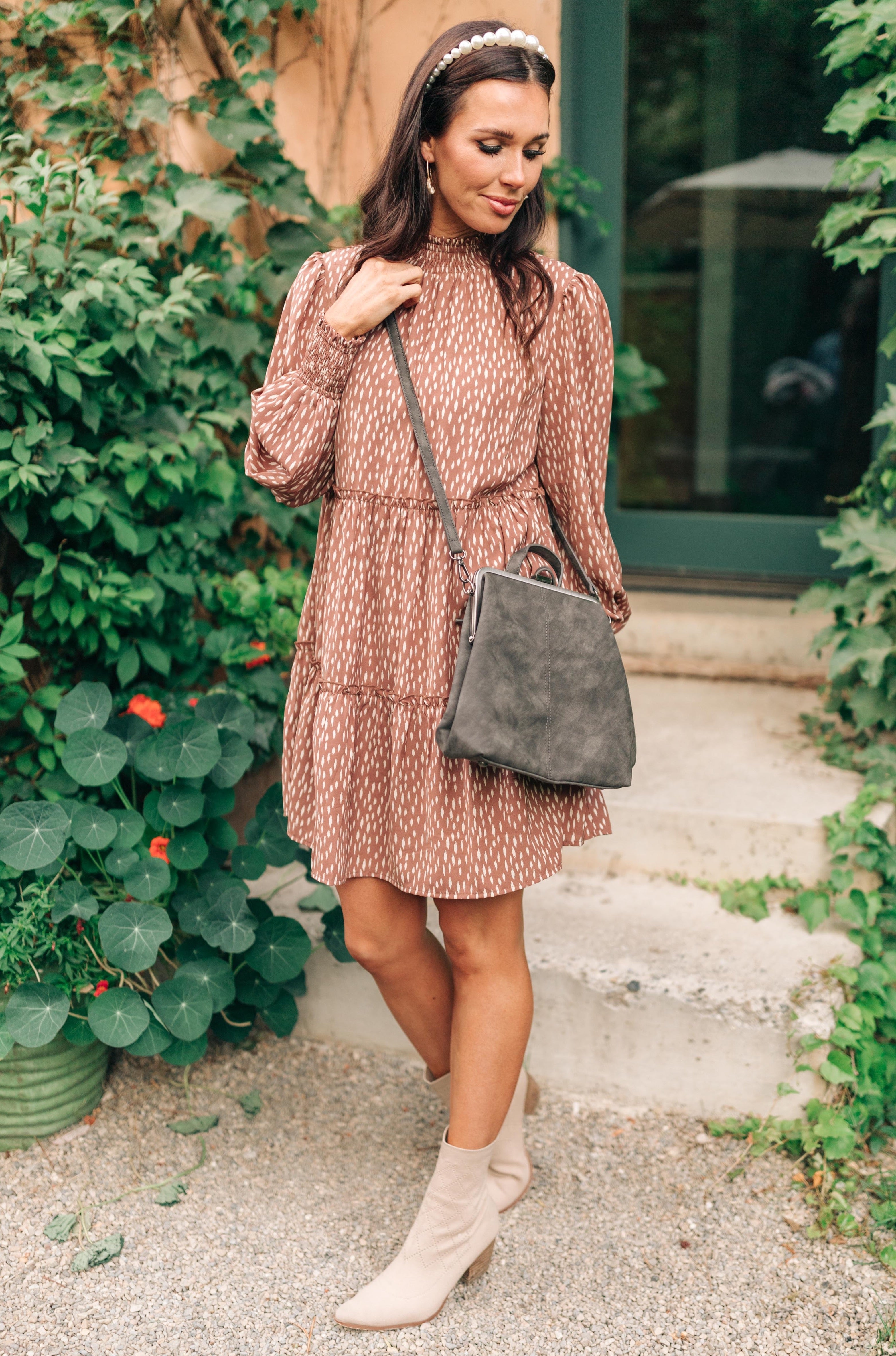 Woman wearing a patterned brown dress and boots carrying a charcoal soft-textured backpack outdoors by greenery.