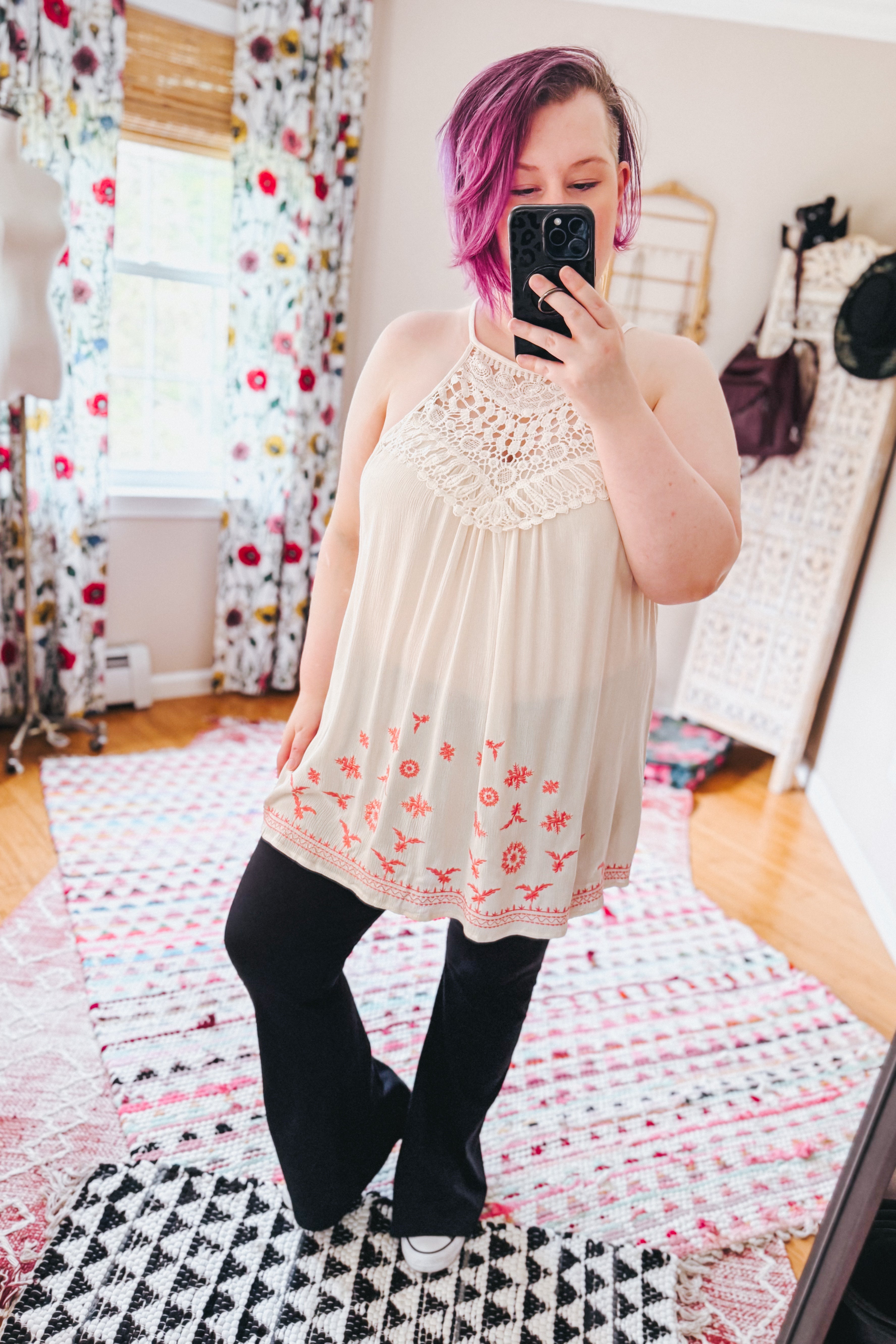 Woman taking mirror selfie wearing cream halter top with crochet yoke and coral embroidery paired with black pants indoors