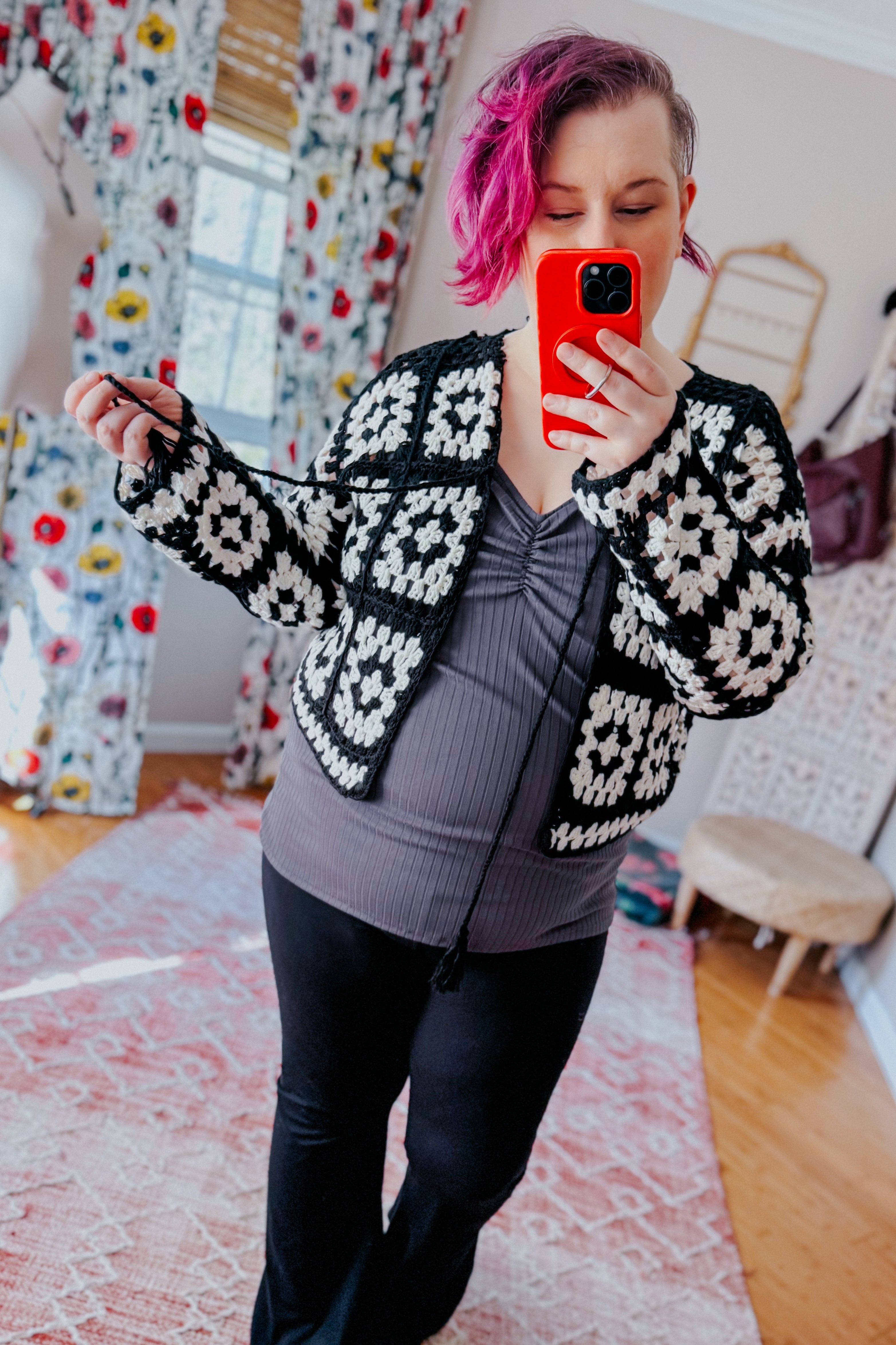 Woman taking mirror selfie wearing black and white crochet cropped cardigan over dark ruched top with black pants indoors