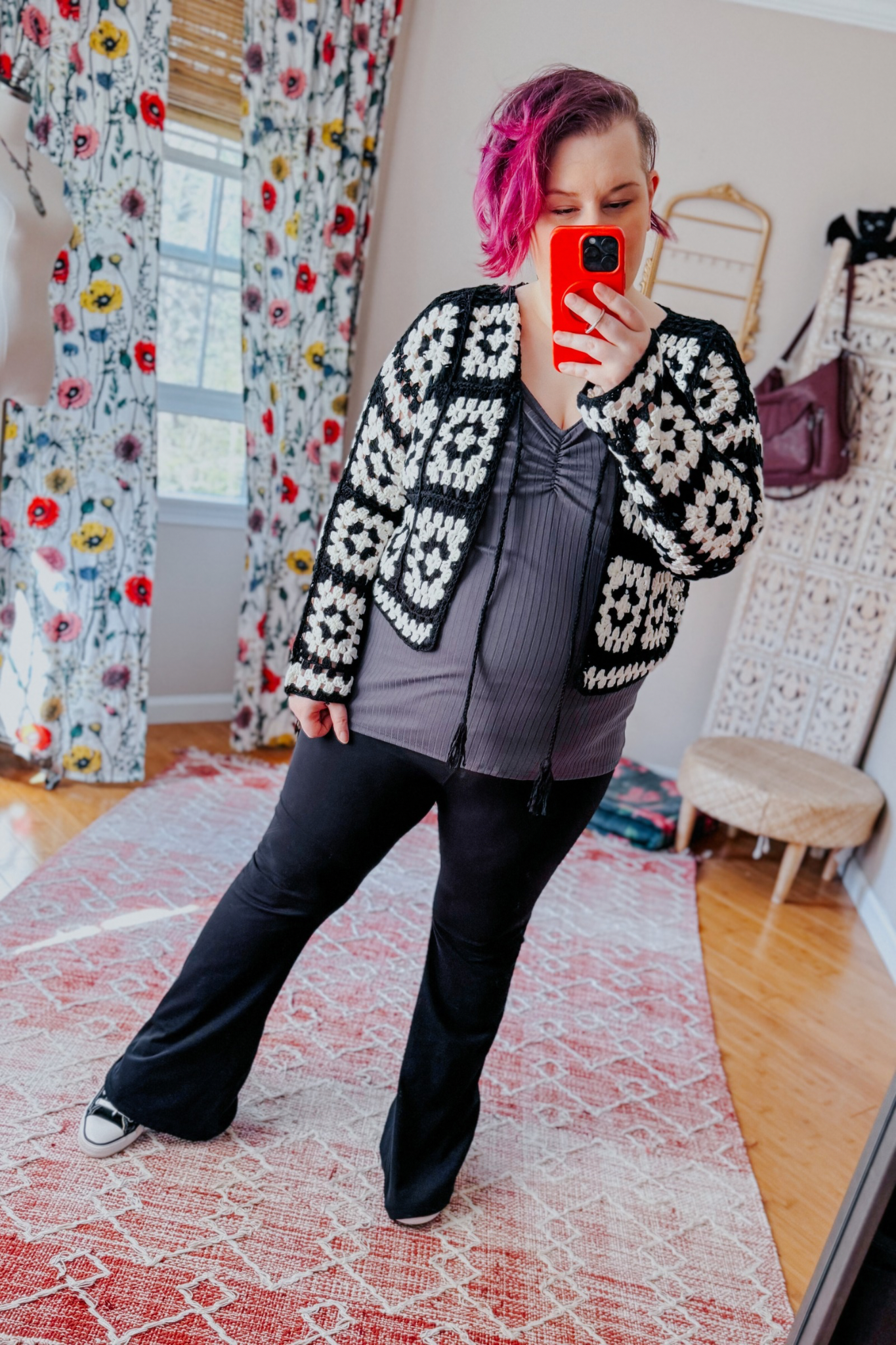 Woman taking indoor mirror selfie wearing black and white crochet cropped cardigan over gray ruched top and black flared pants