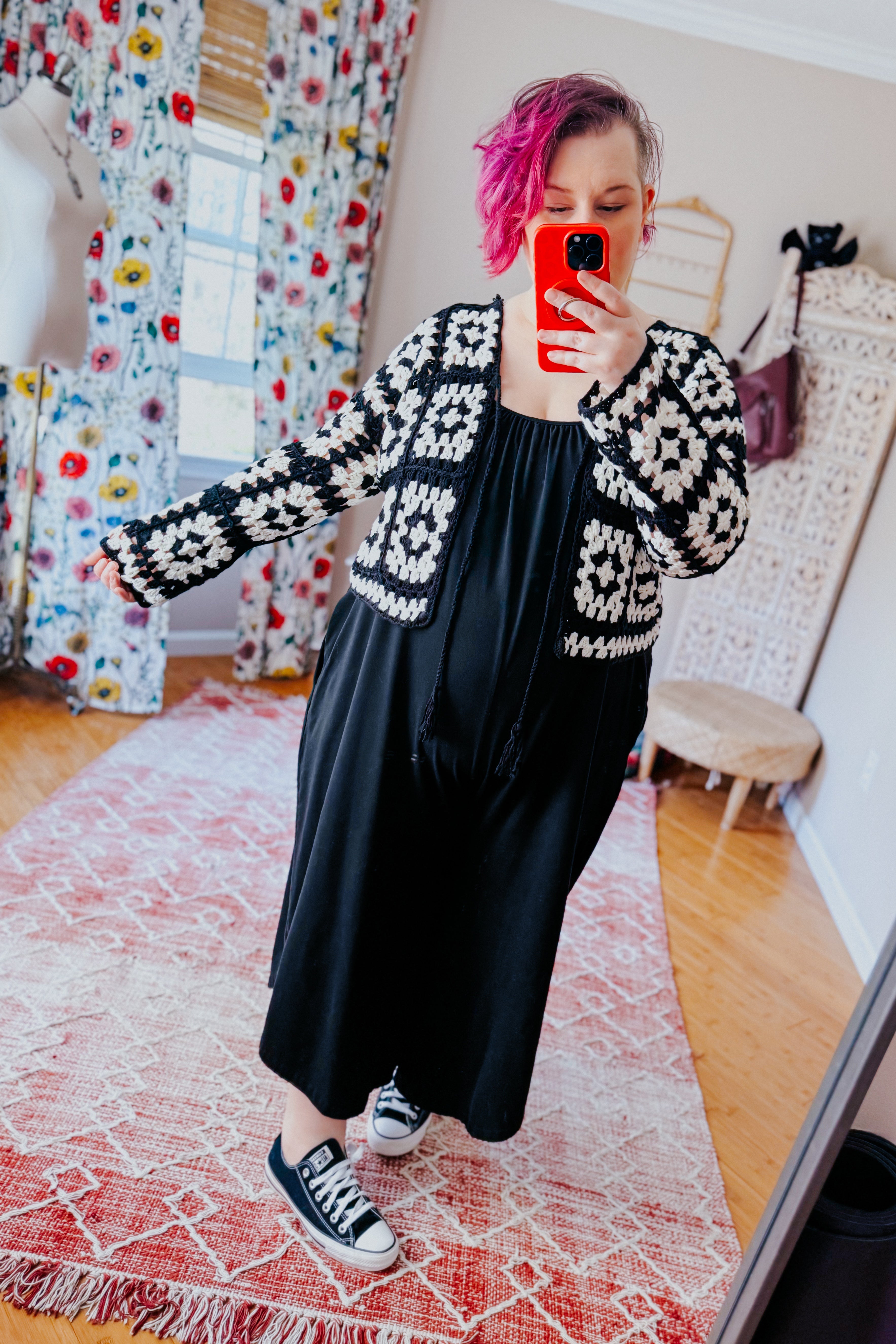 Woman indoors taking mirror selfie wearing black dress with black and white crochet patterned cropped cardigan and sneakers