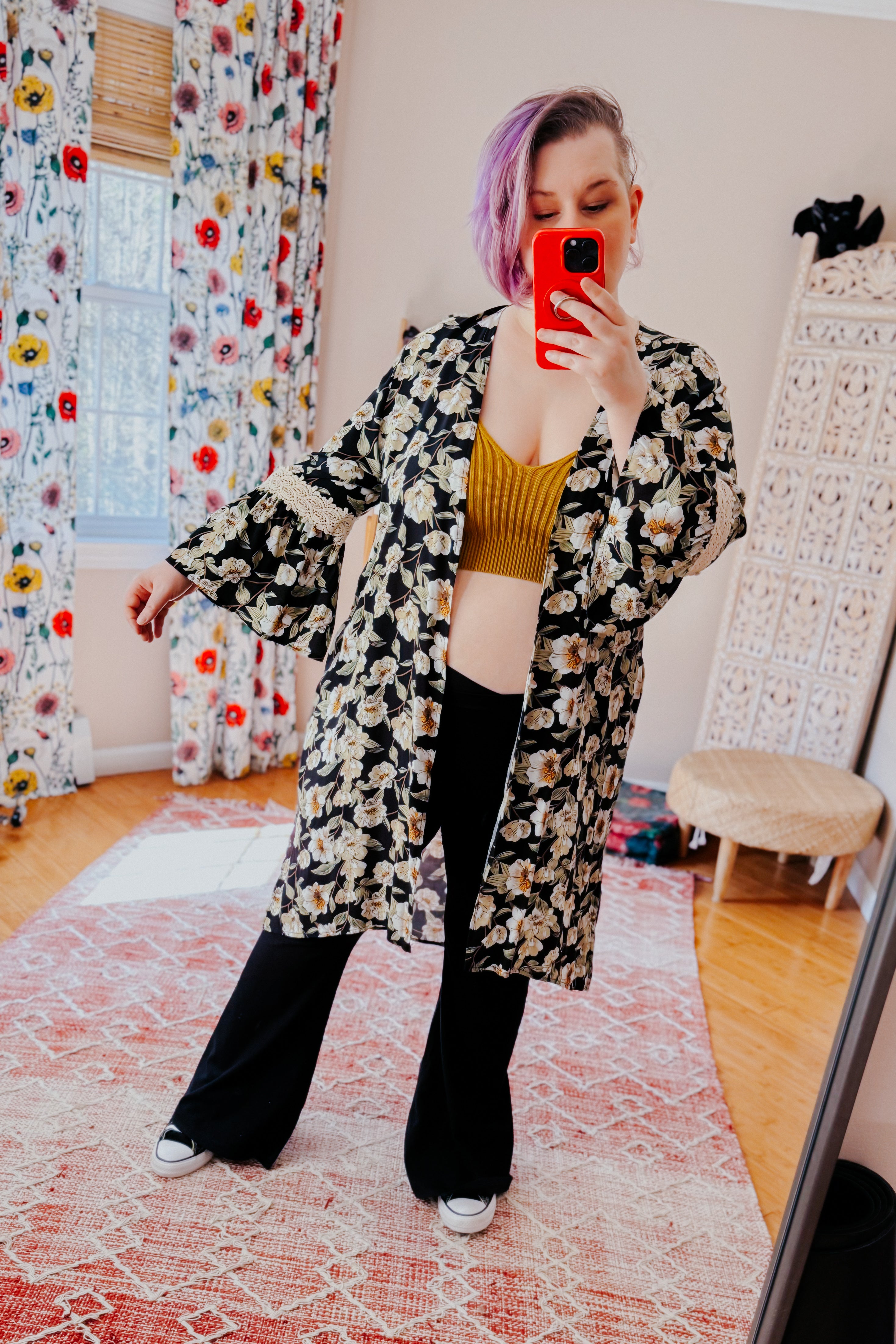 Woman with short purple hair wearing black floral 3/4 bell sleeve duster over mustard ribbed top and black pants indoors taking selfie