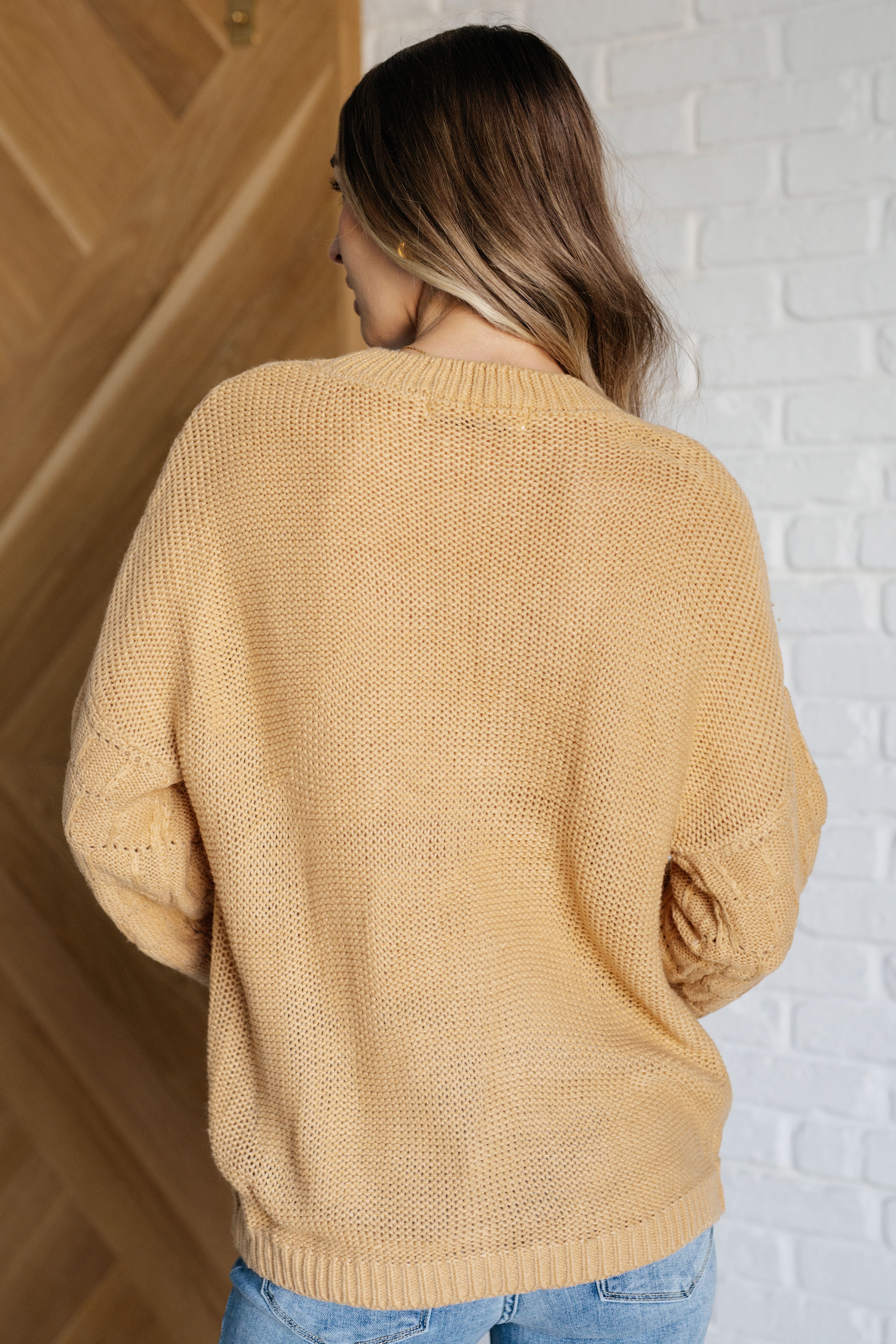Woman wearing wheat-colored bubble sleeve sweater facing away against white brick and wooden door background