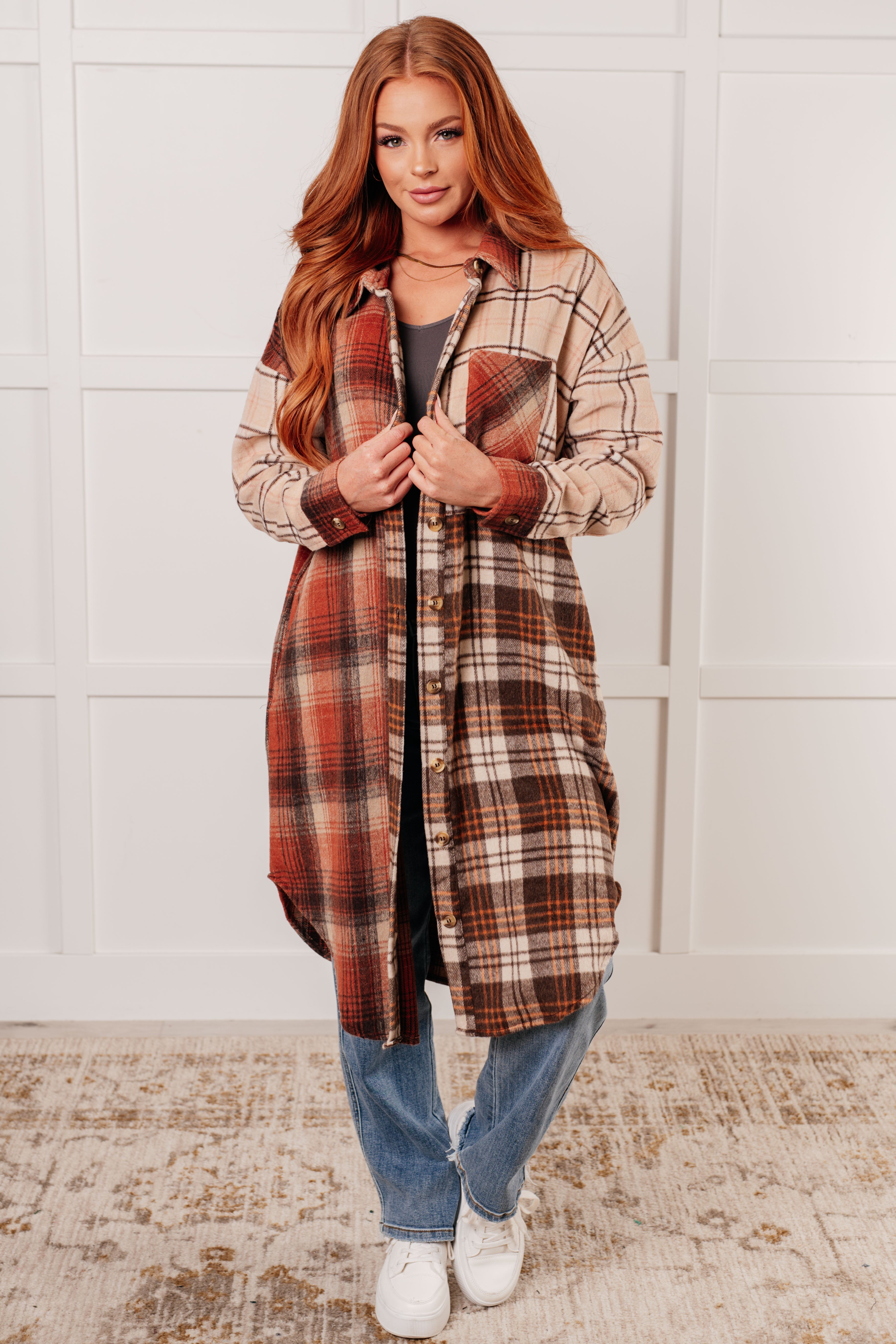 Woman wearing a long plaid flannel shacket in cream, rust, and brown tones with blue jeans and white sneakers indoors