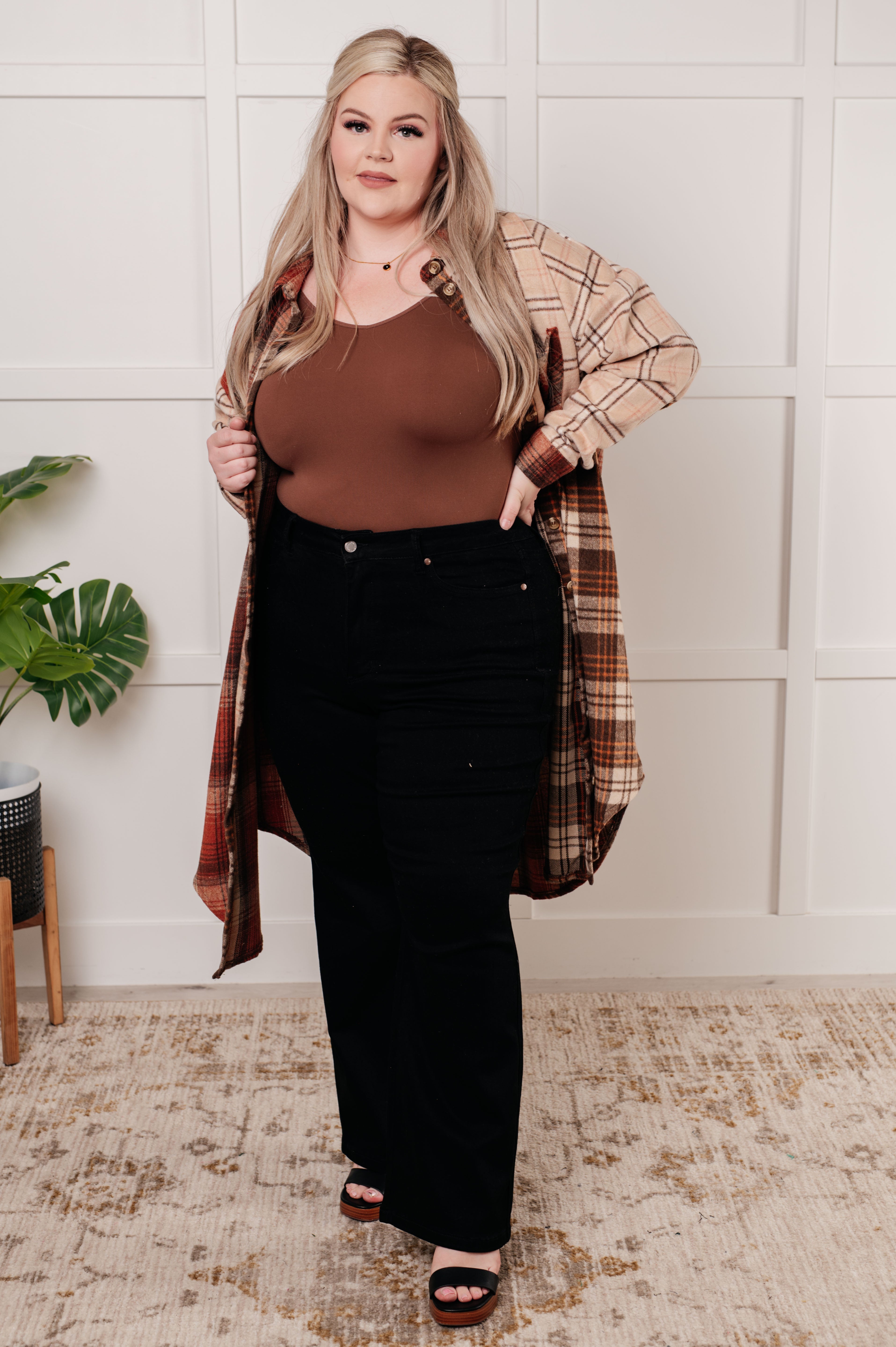 Plus-size woman wearing beige and brown plaid oversized flannel shacket with brown tank top and black pants indoors