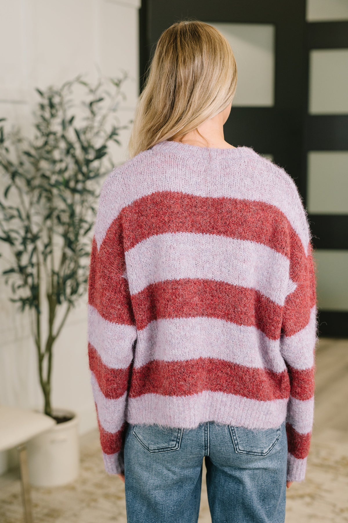 Back view of a woman wearing a red and purple striped drop shoulder sweater with relaxed sleeves and blue jeans indoors