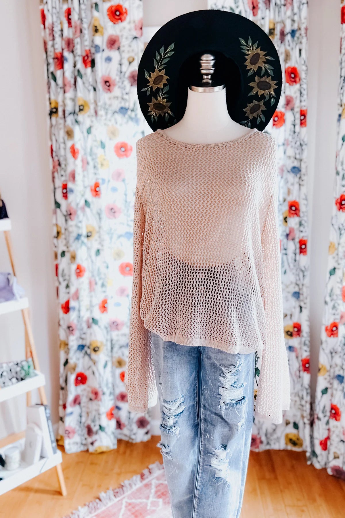 Beige loose knit long sleeve top styled on mannequin with distressed light blue jeans and black floral wide-brim hat indoors