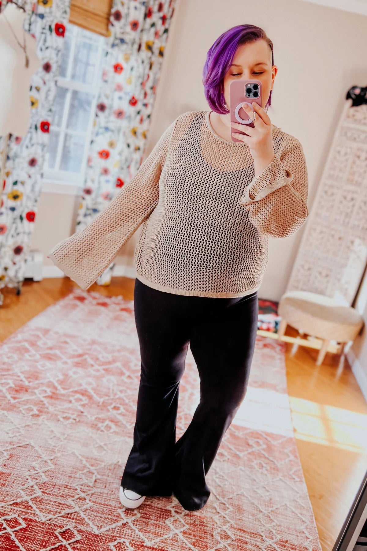 Woman with purple hair wearing beige loose knit top and black flared pants taking indoor mirror selfie on patterned rug
