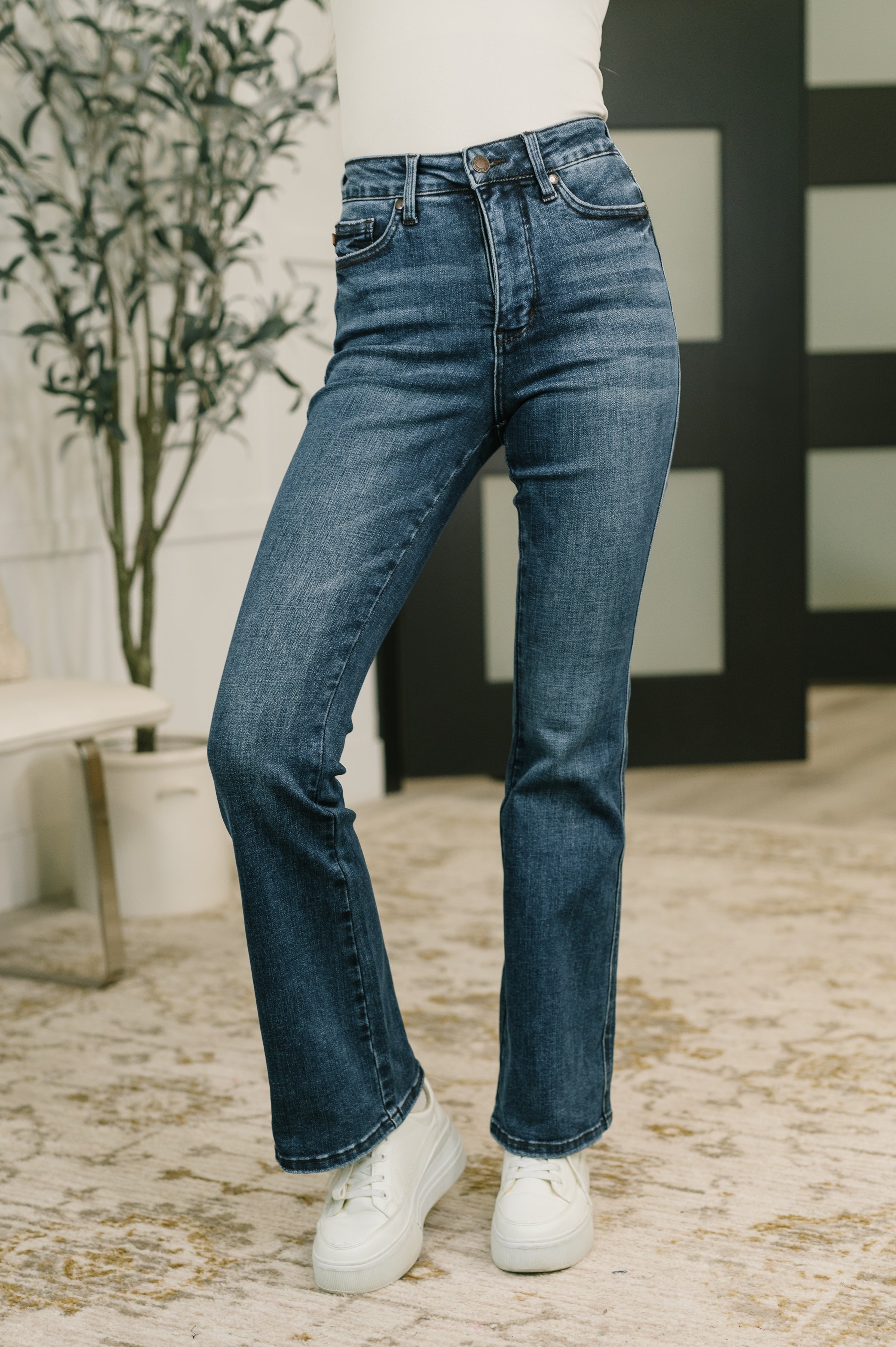 Woman modeling blue high waist bootcut jeans with subtle fading and white sneakers in a bright indoor setting