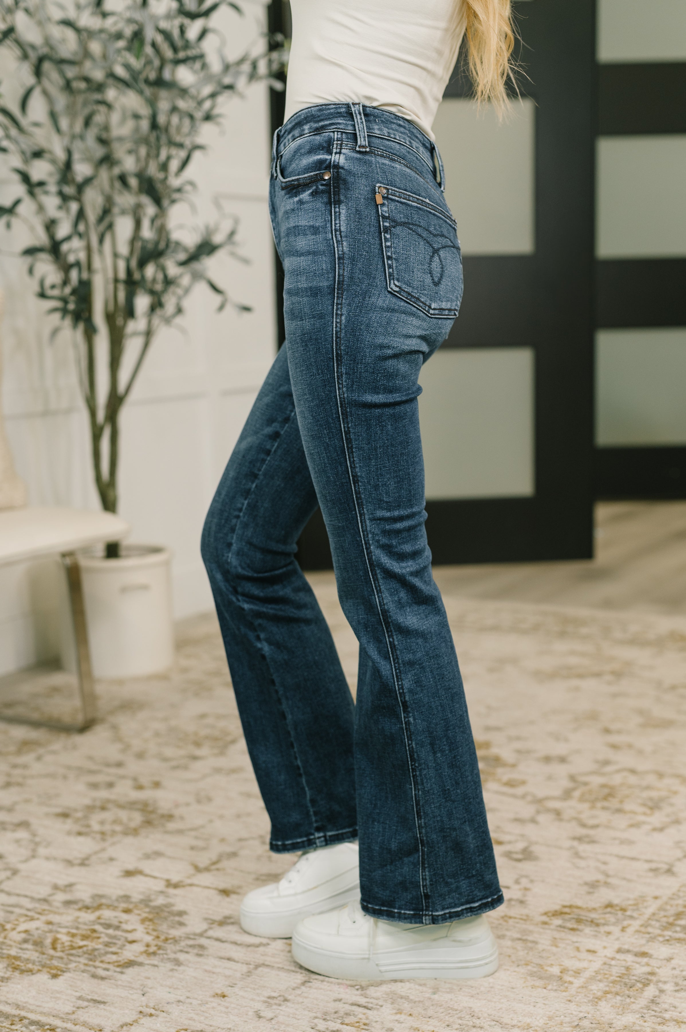 Side view of a woman wearing dark wash high waist bootcut jeans with a white top and sneakers in a bright indoor room