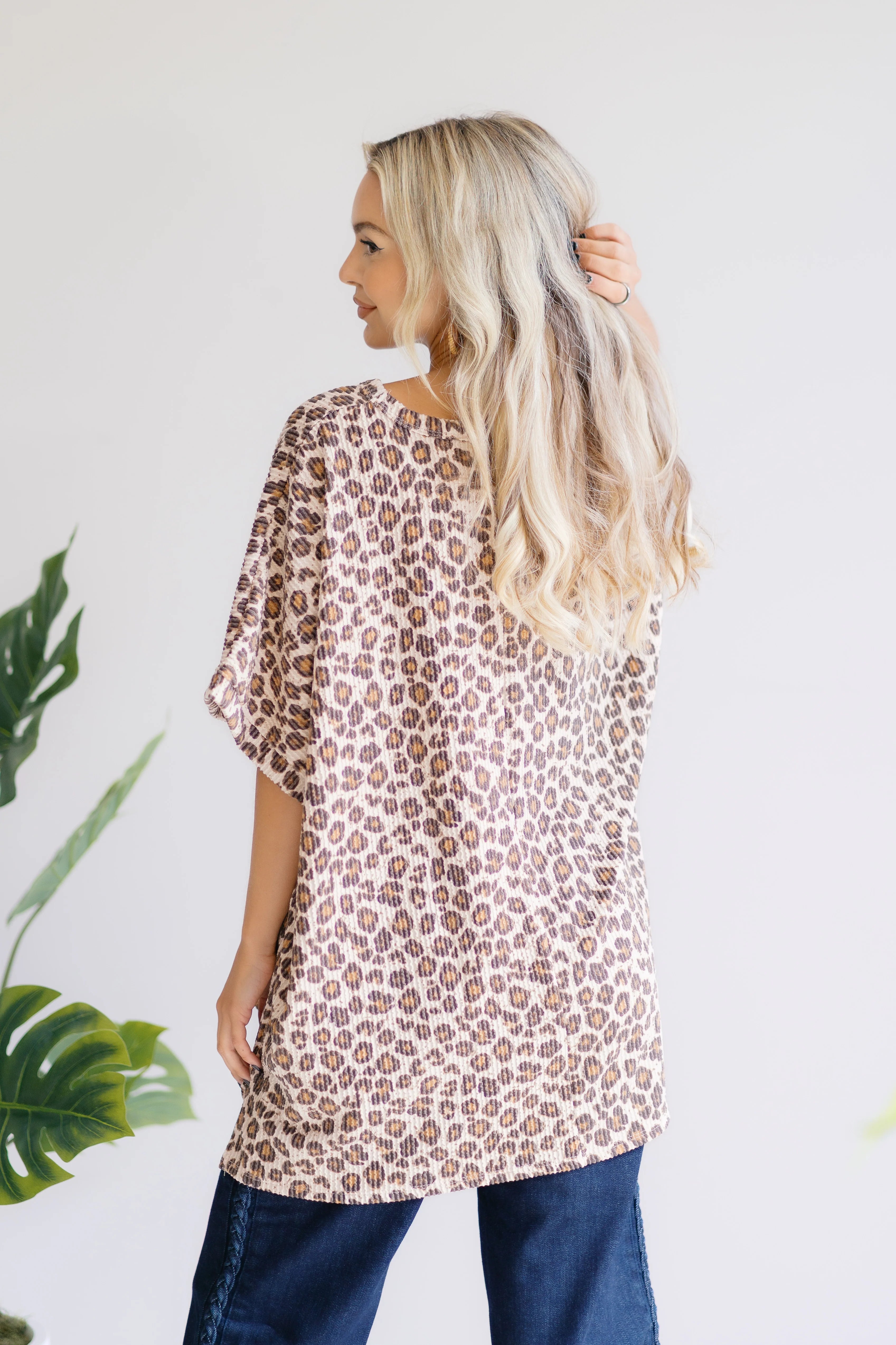 Blonde woman with wavy hair posing in loose leopard print Canyon Wild Top and dark blue jeans against white background