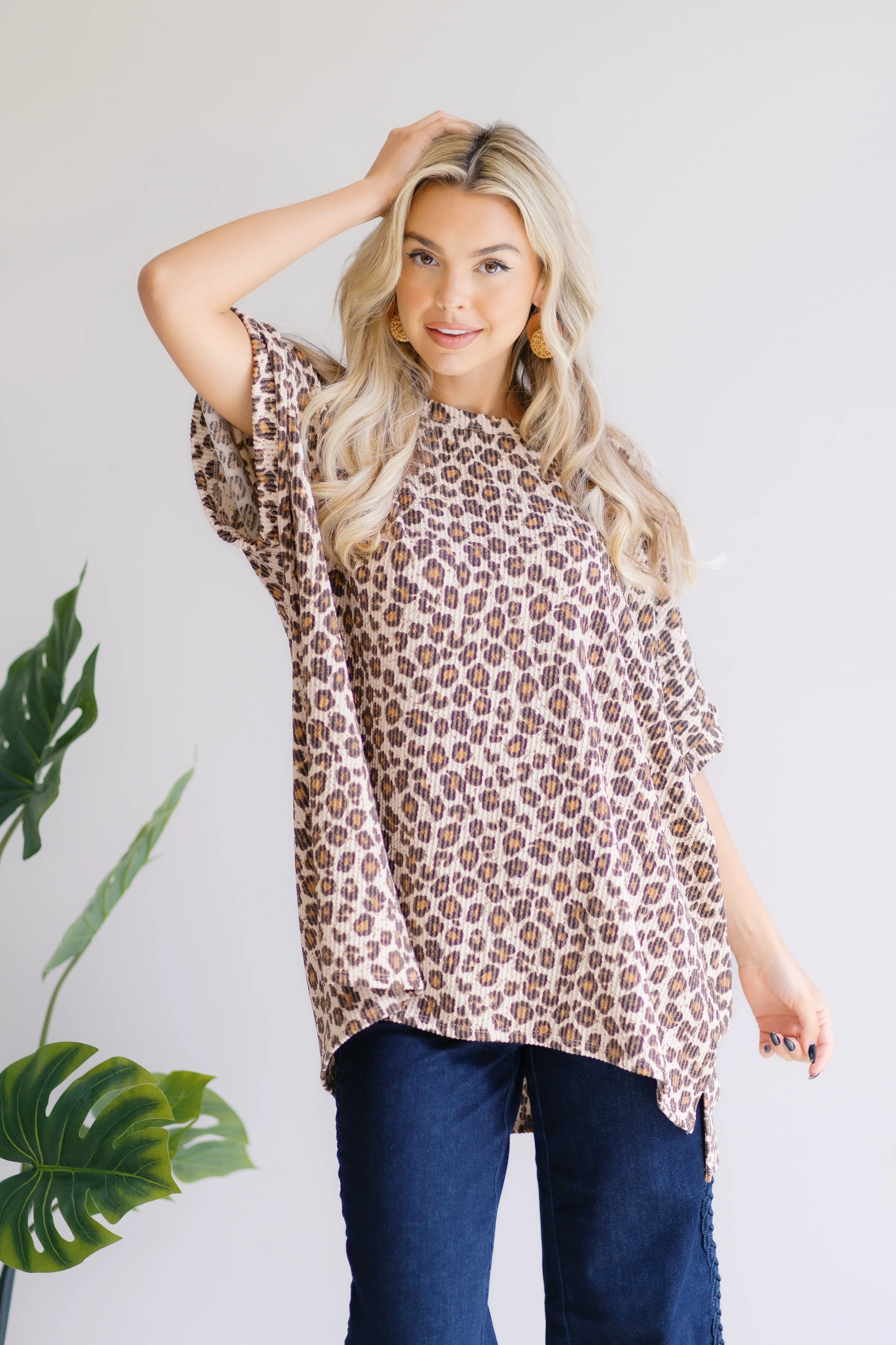 Blonde woman wearing loose leopard print Canyon Wild Top with dark blue jeans posing next to green plant indoors