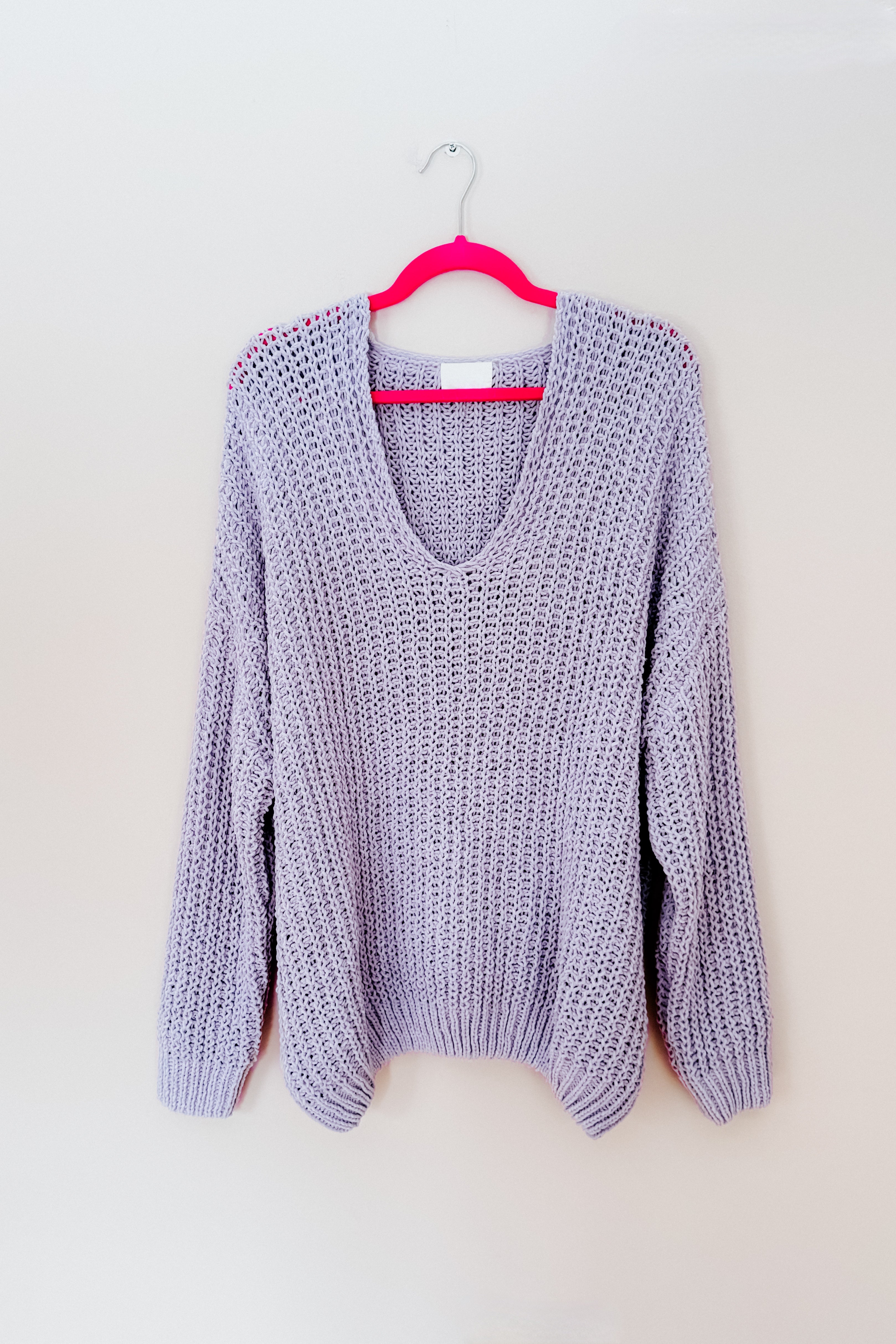 Light purple chunky knit V-neck sweater displayed on a bright pink hanger against a plain wall
