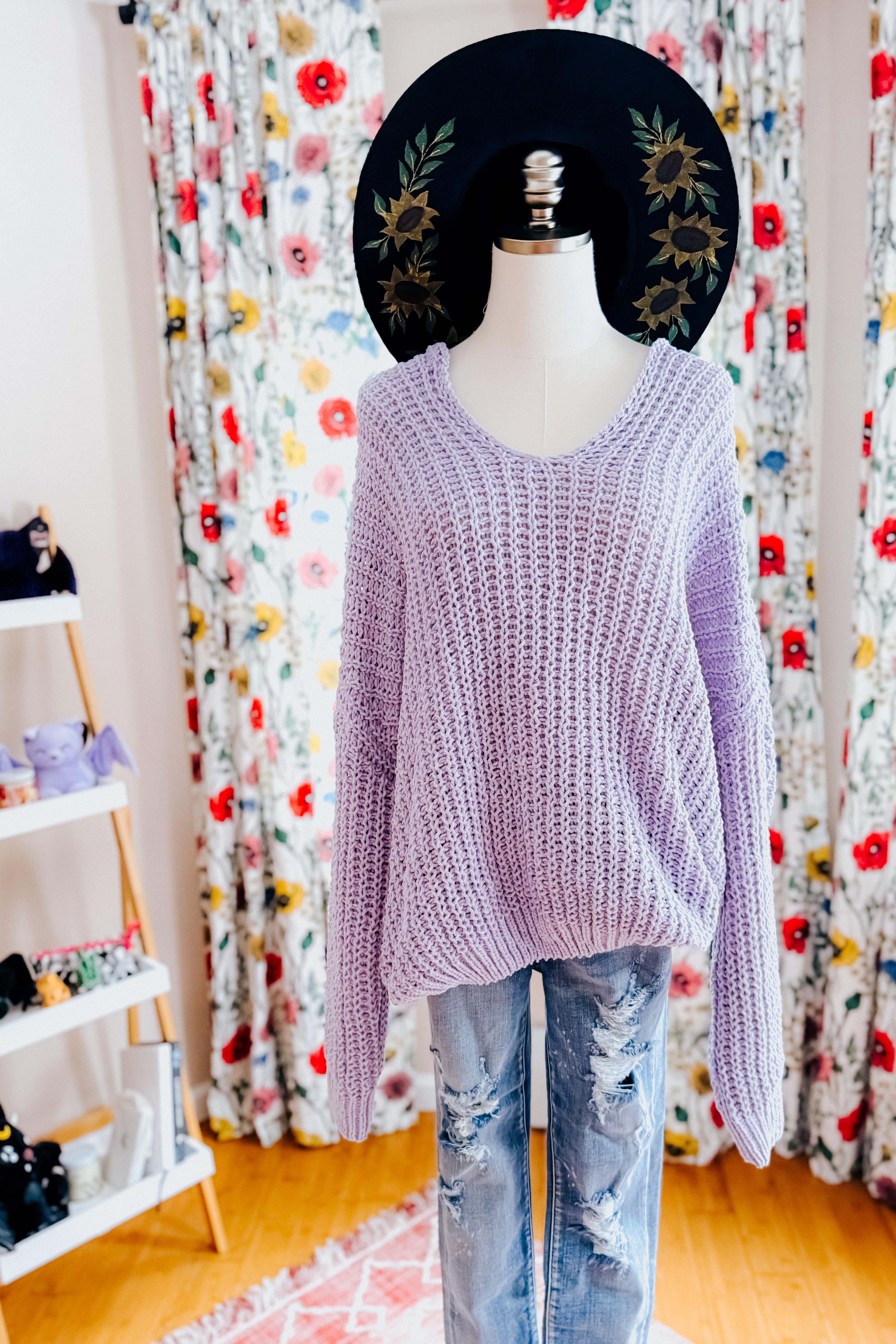 Loose chunky lavender V-neck sweater styled on a mannequin with distressed light blue jeans and a black floral wide-brim hat indoors