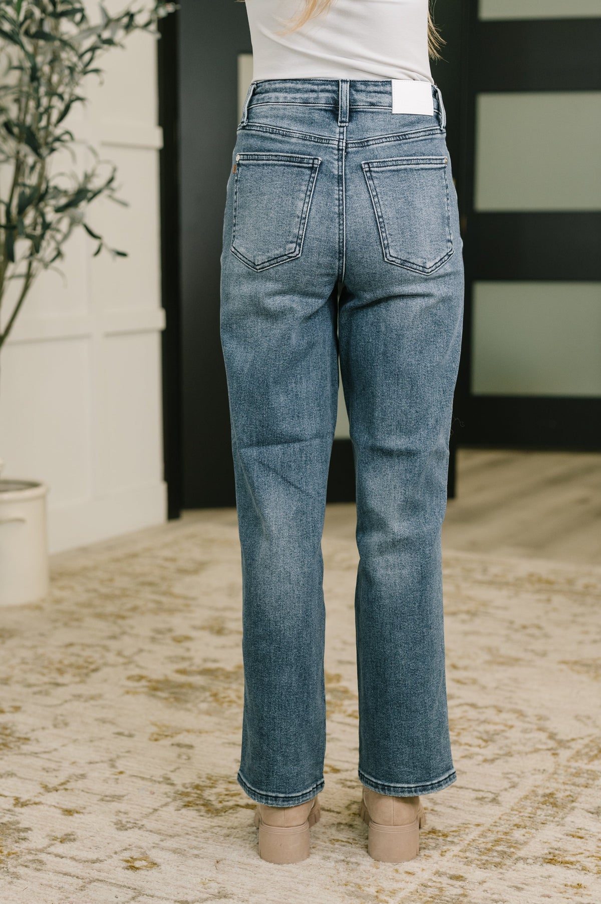 Back view of a woman wearing blue high waist straight leg jeans with back pockets and a white top styled with neutral shoes indoors