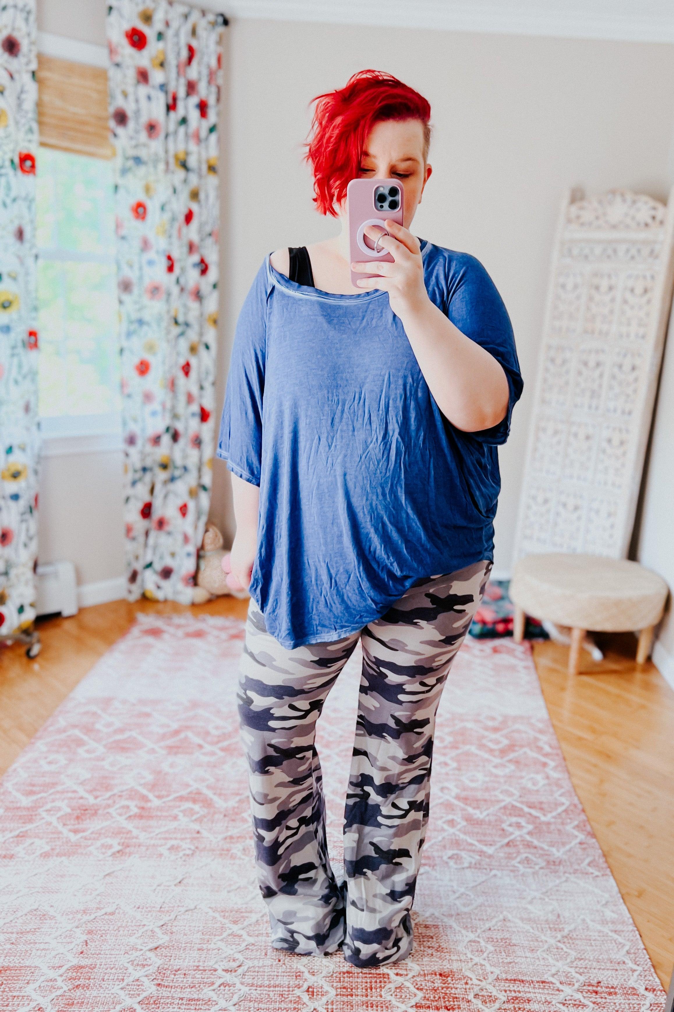 Woman taking mirror selfie wearing loose blue top and light grey and navy blue camouflage flare pants in cozy living space