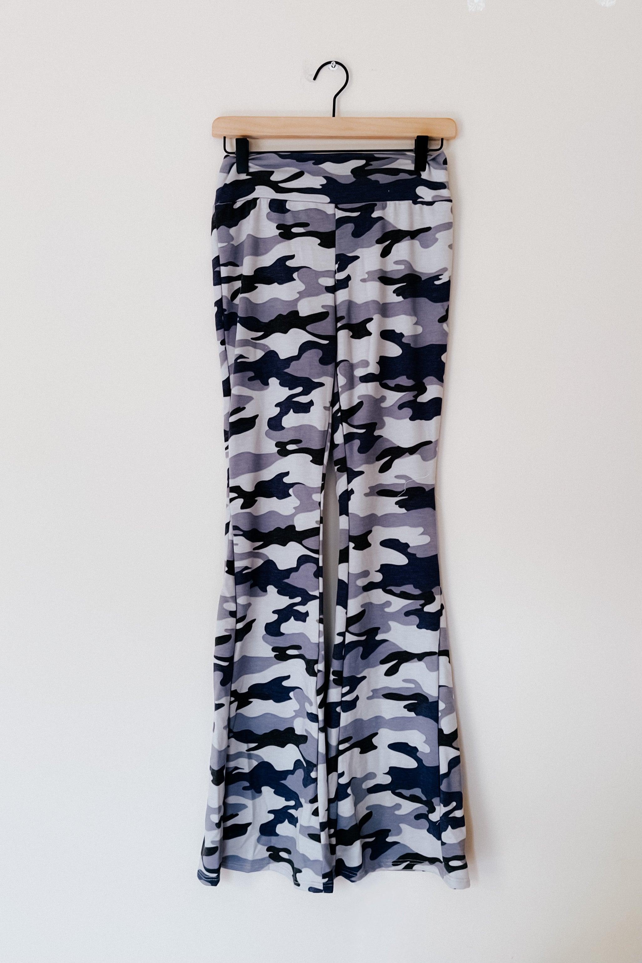 Gray and navy blue camouflage flare pants hanging on wooden hanger against white wall background
