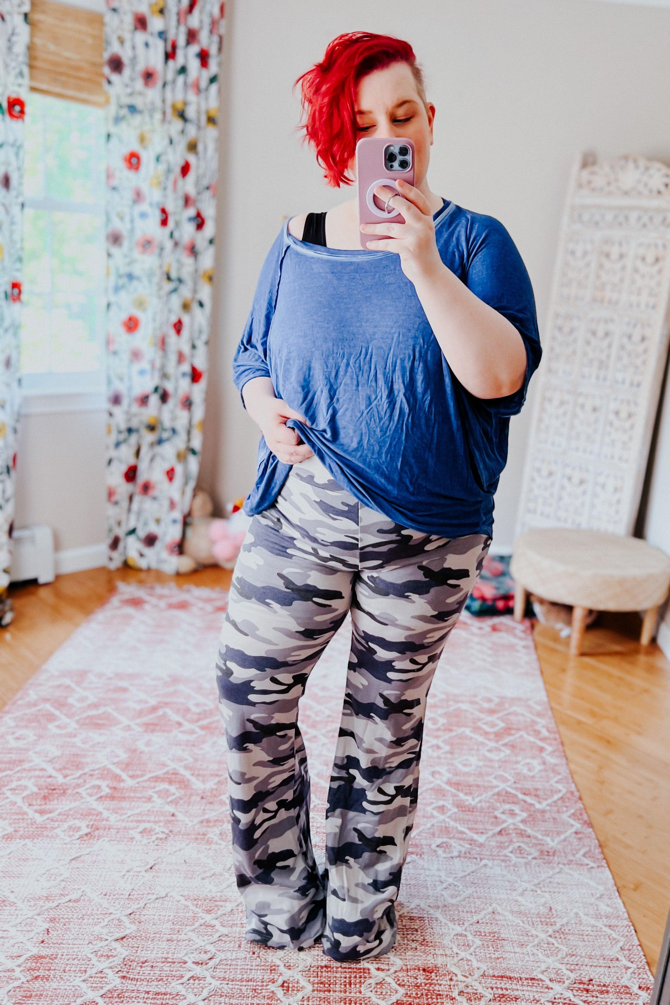 Plus size model wearing light gray and navy blue camouflage flare pants with loose blue top taking mirror selfie indoors
