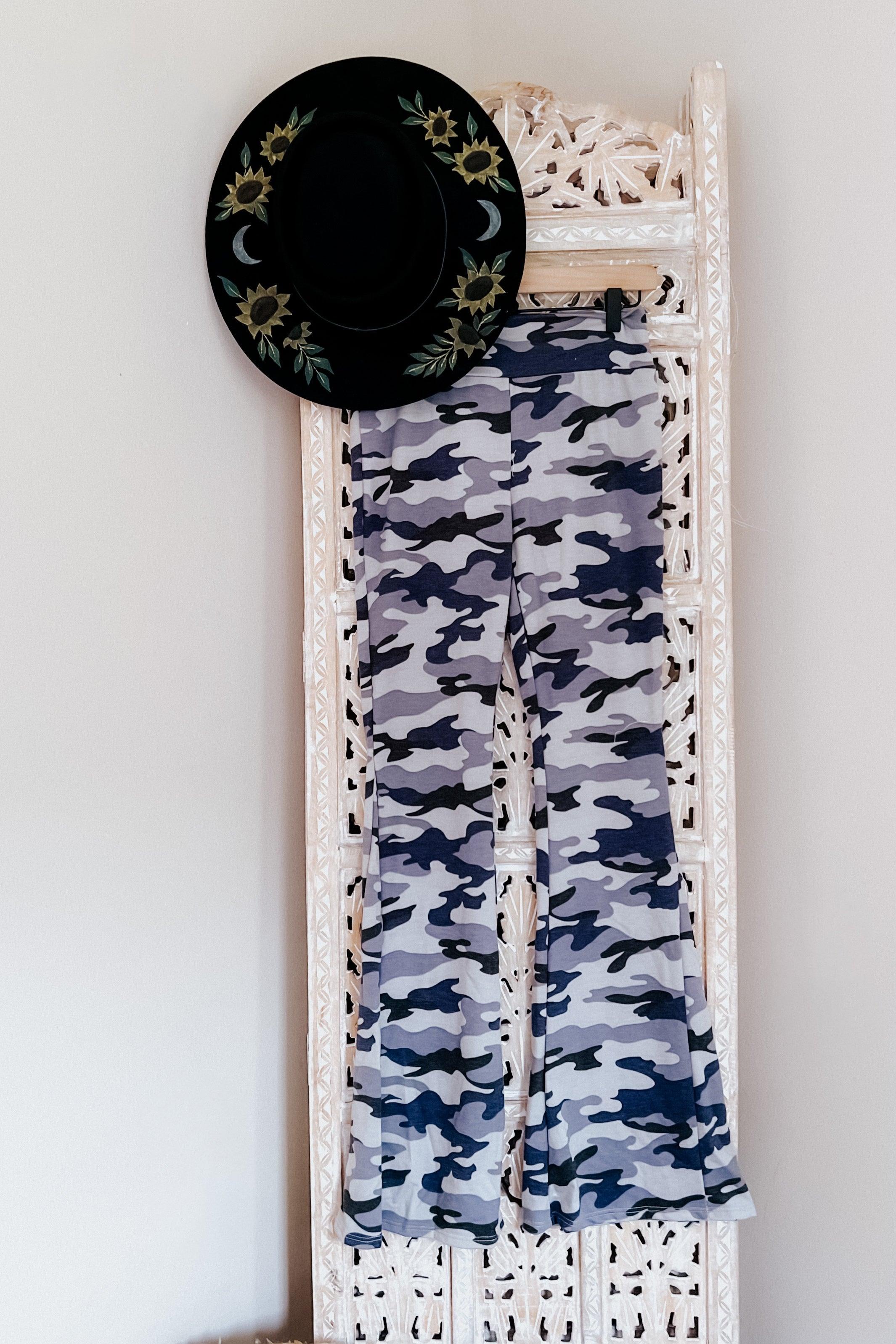 Light gray and navy blue camo flare pants hanging on carved white wooden panel with black embroidered hat