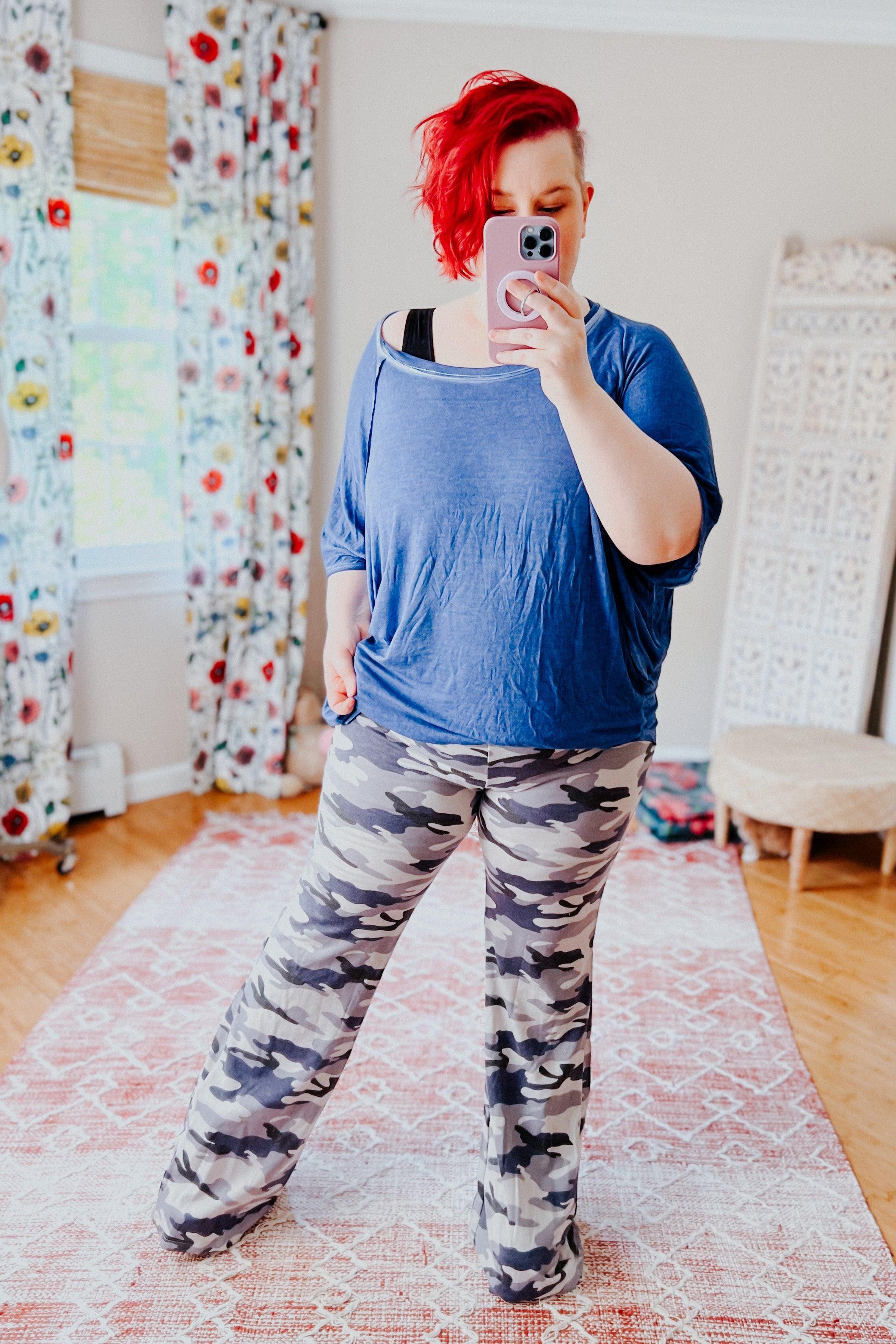 Woman wearing loose blue top and light gray and navy blue camo flare pants taking mirror selfie in bright room with floral curtains
