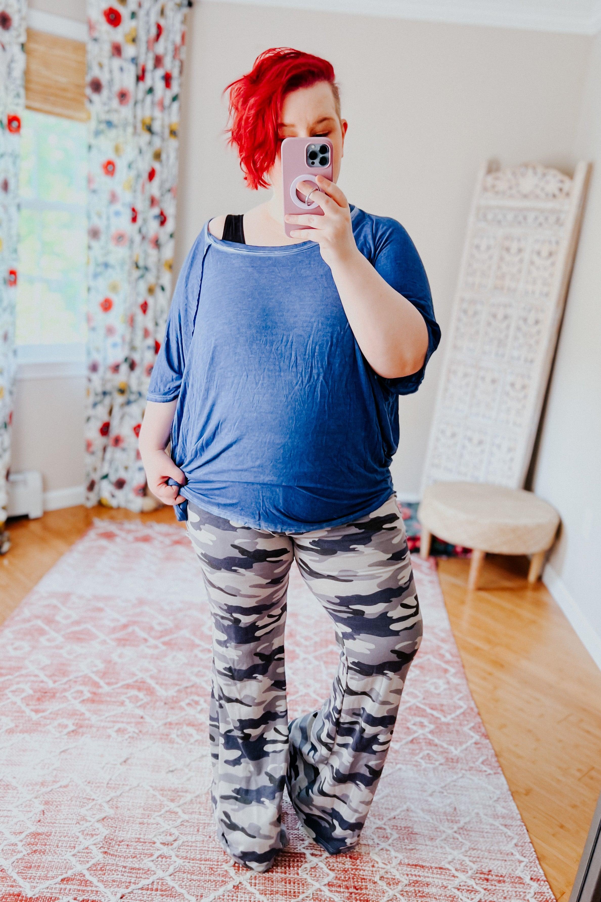 Woman with red hair wearing loose blue top and grey and navy blue camo flare pants taking selfie indoors on patterned rug