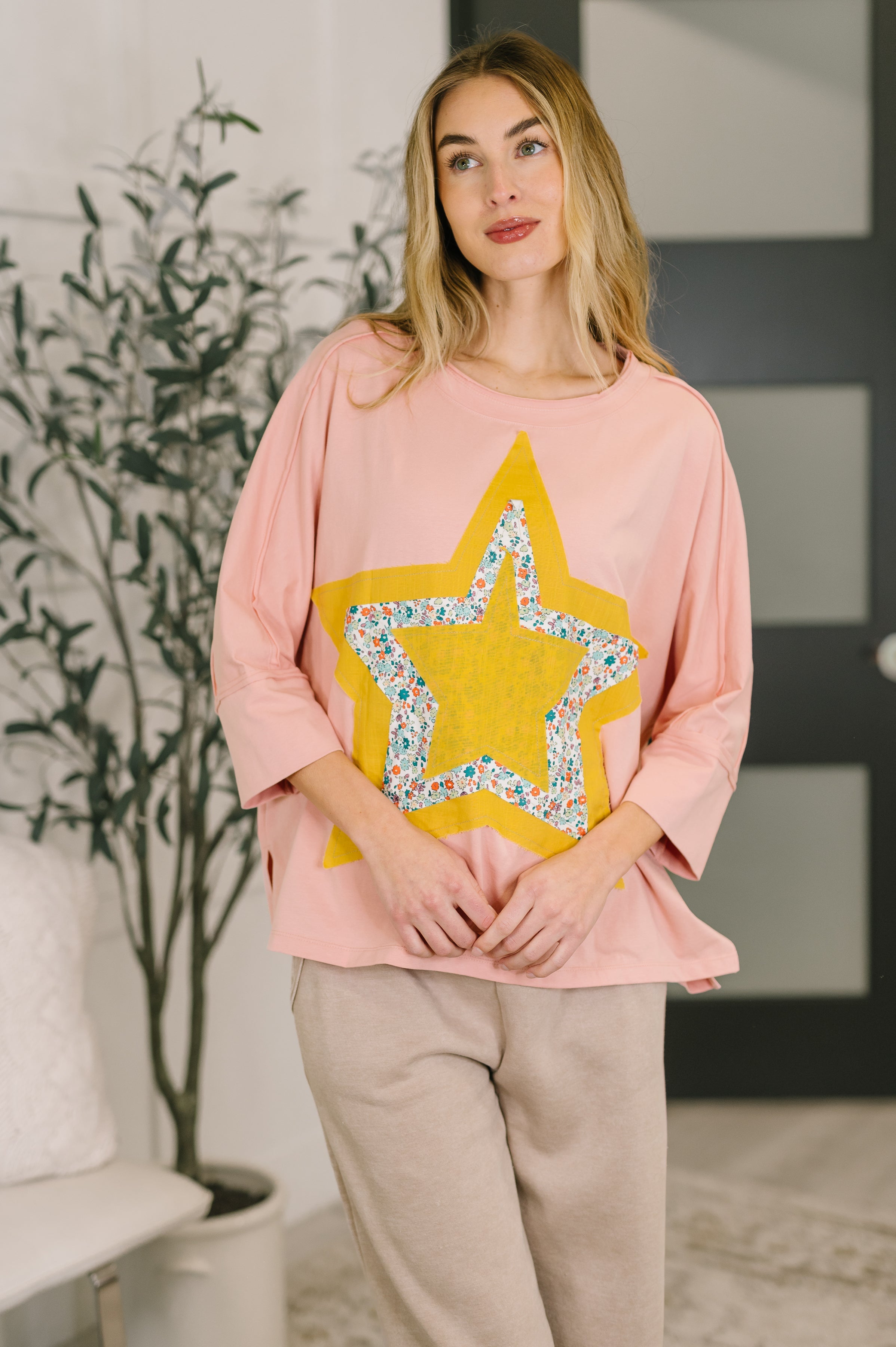 Woman wearing loose pastel pink top with large yellow star and floral mixed print star patch paired with beige pants indoors