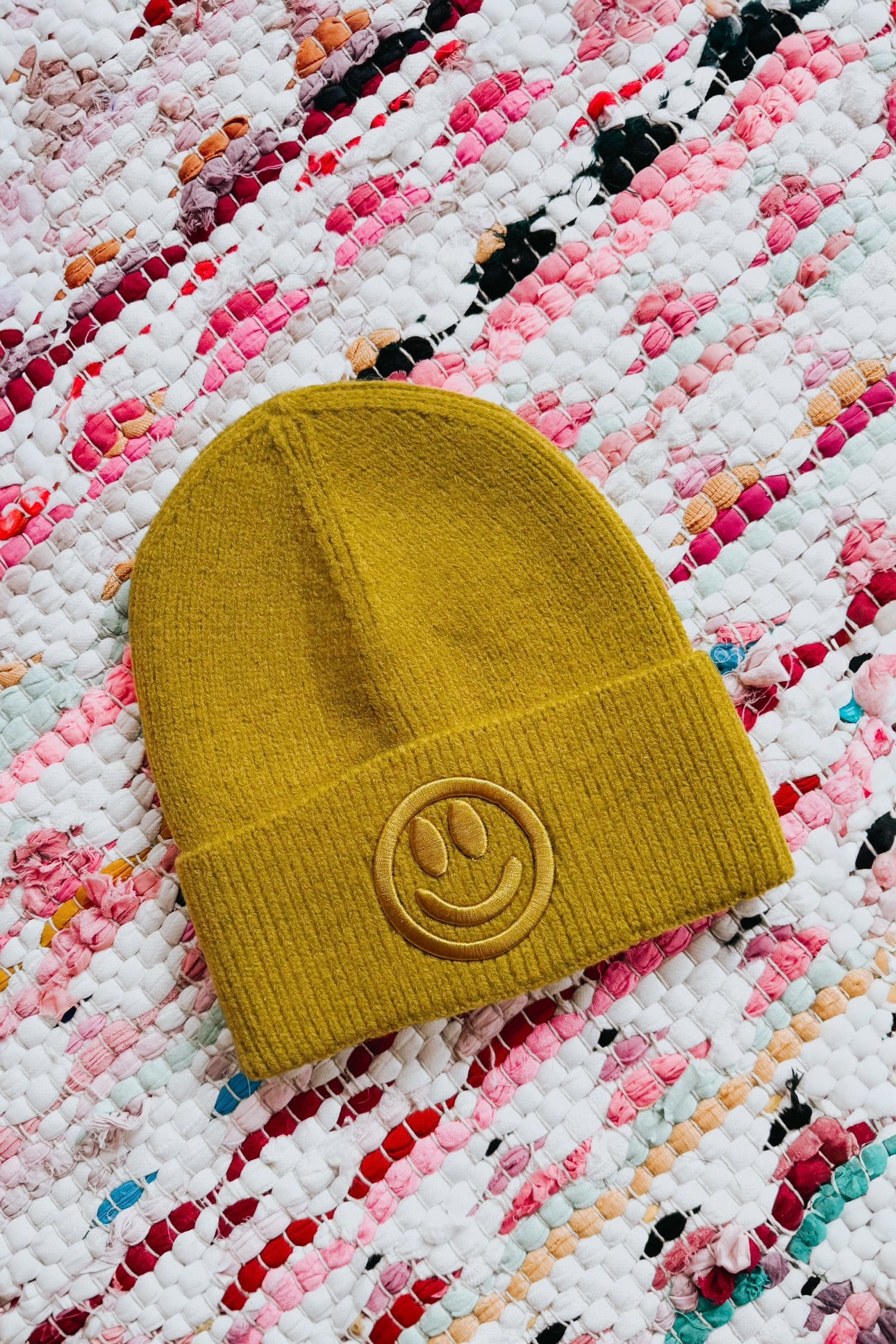 Mustard yellow ribbed knit beanie with embroidered smiley face emblem lying on colorful woven fabric background