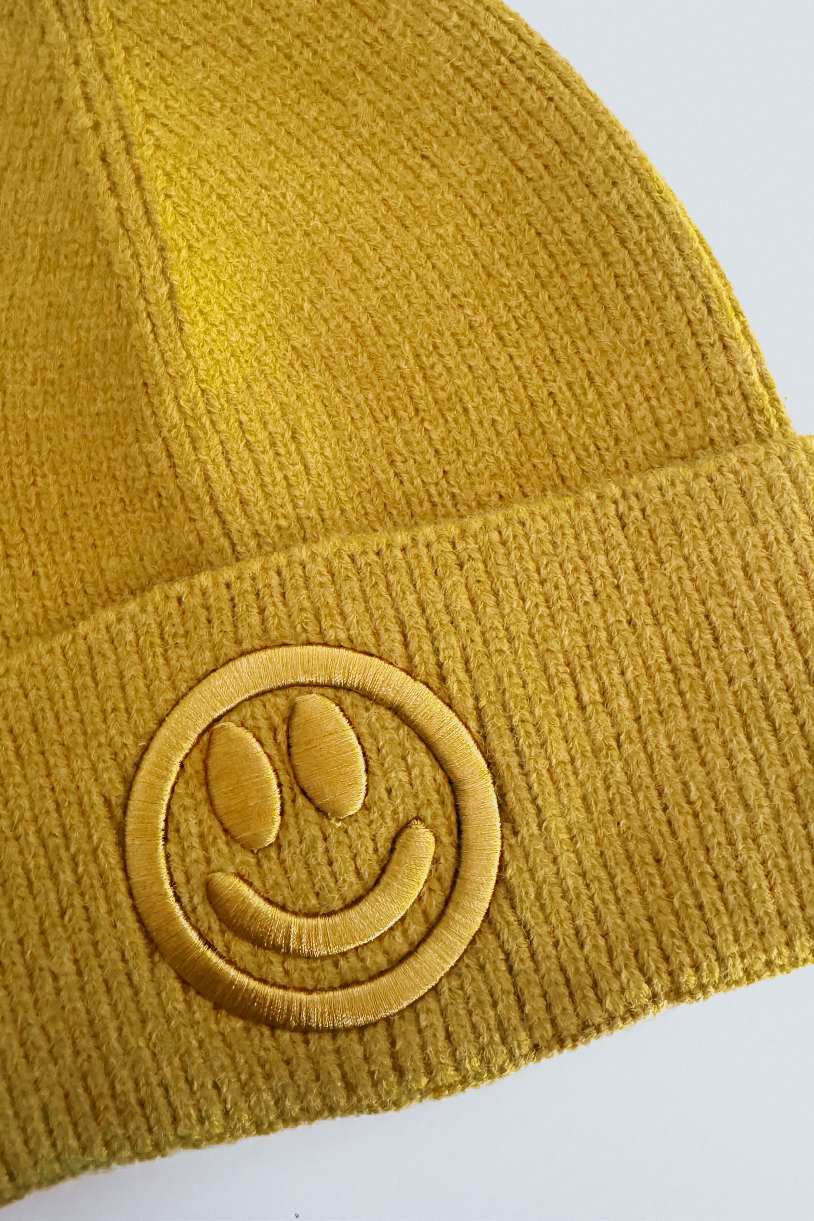 Close-up of mustard yellow ribbed knit beanie with raised embroidered smiley face patch on cuff against plain background