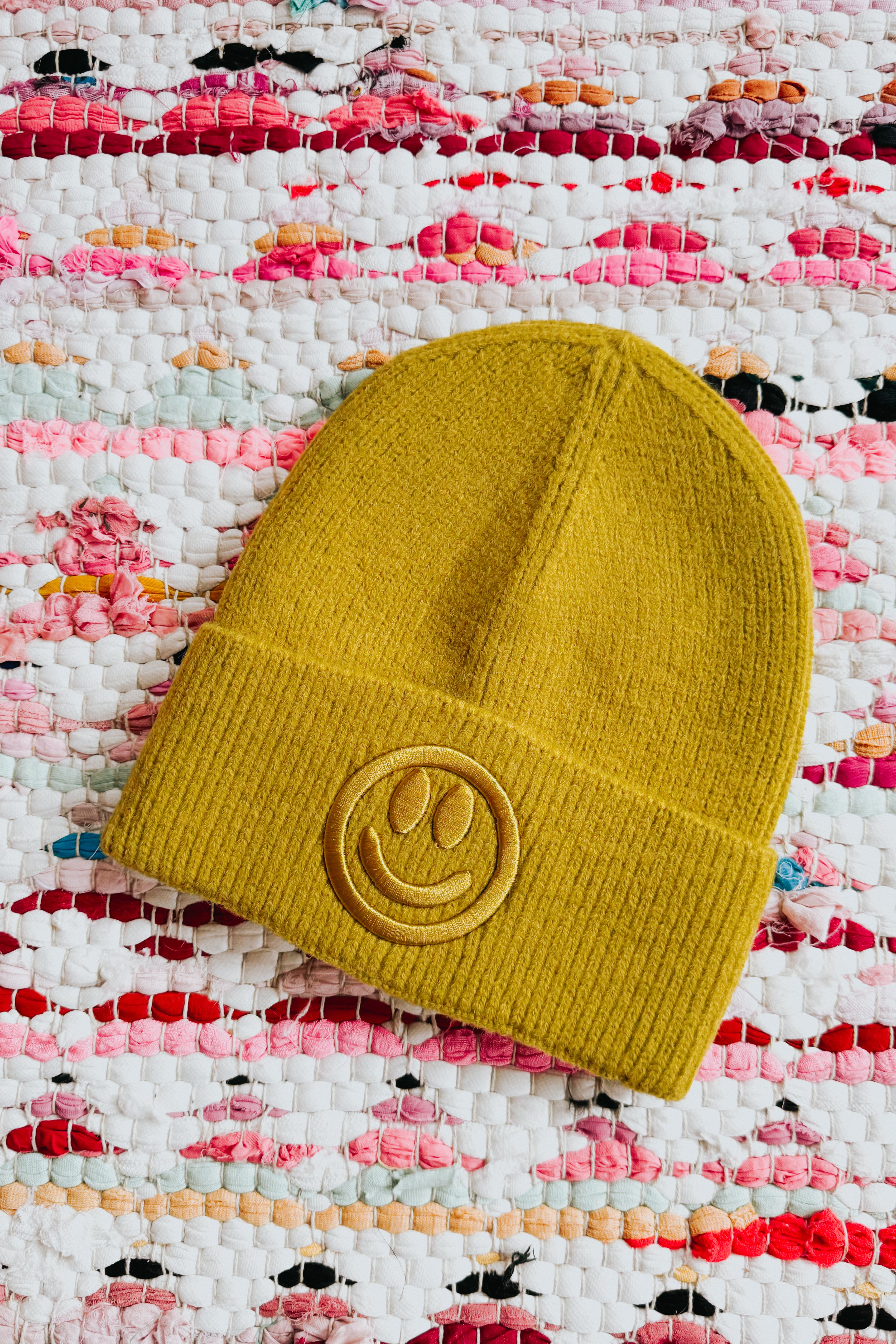 Mustard yellow ribbed knit beanie with embroidered smiley face lying on woven multicolor fabric background